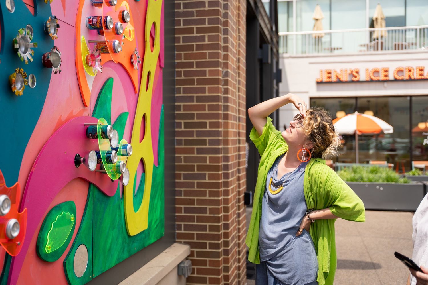 A person in a lime green cardigan views a vibrant colorful wall mural outside a brick building.
