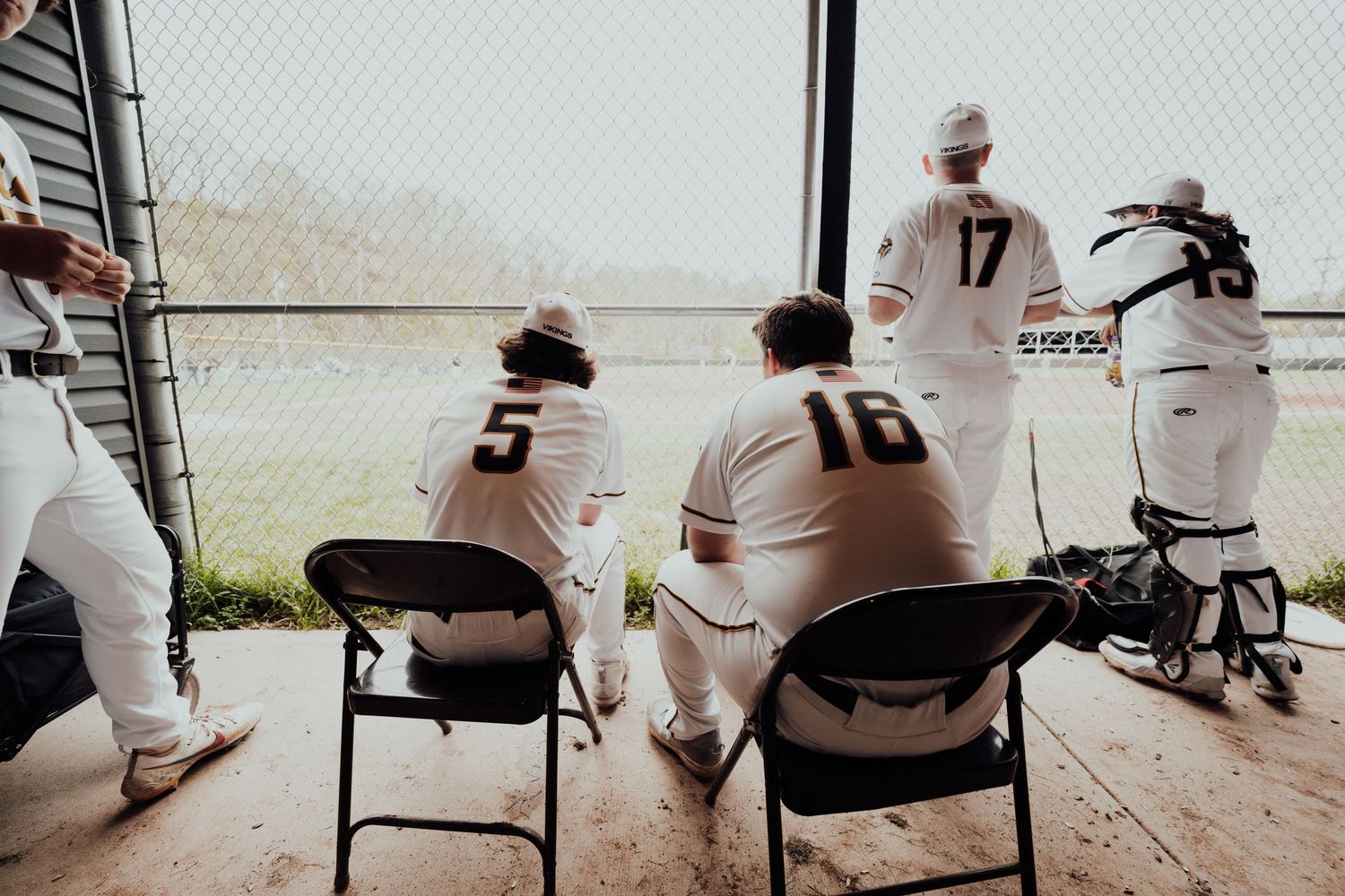 Baseball players and coaches sit in dugout chairs watching the game through chain link fence.