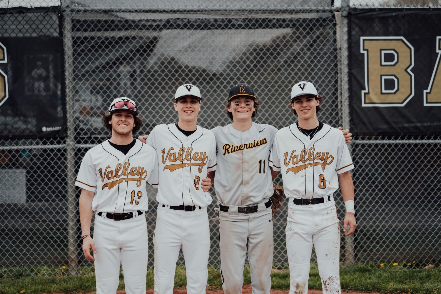 Baseball team wearing white jerseys poses together in front of a chain link fence.