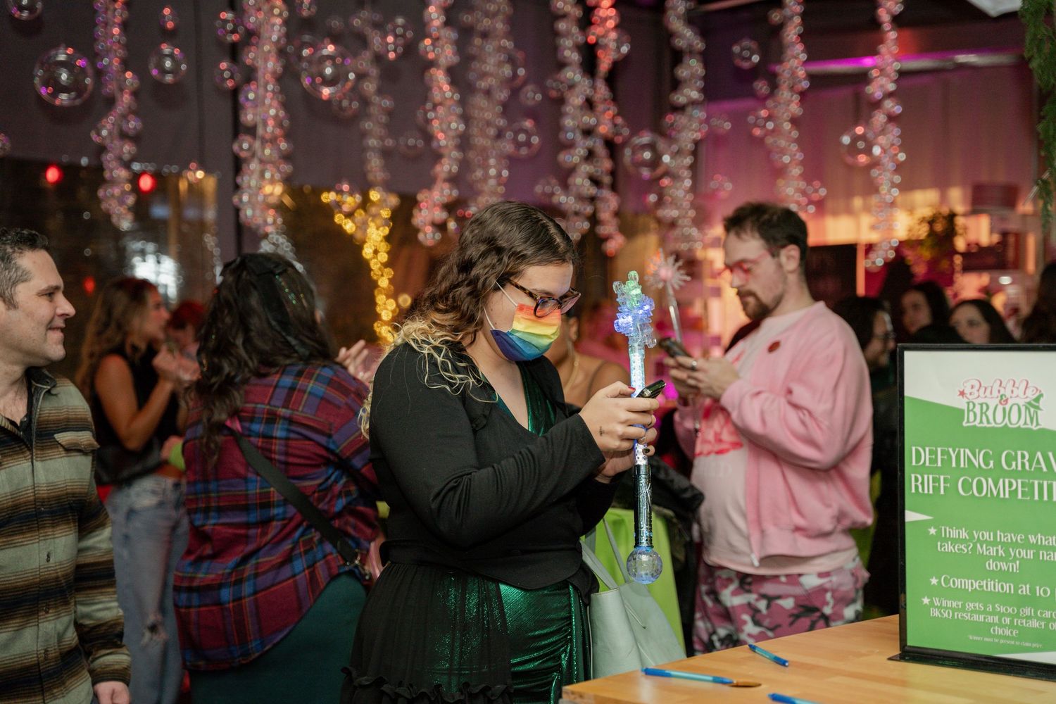 Indoor party scene with festive string lights and bubbles while someone in a face mask holding a blue drink.