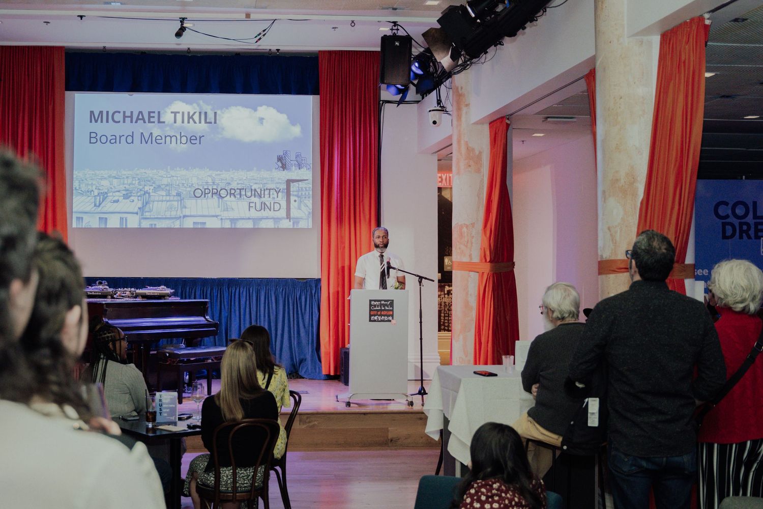 People gathered in a dimly lit venue with red curtains and projection screens for an event presentation.