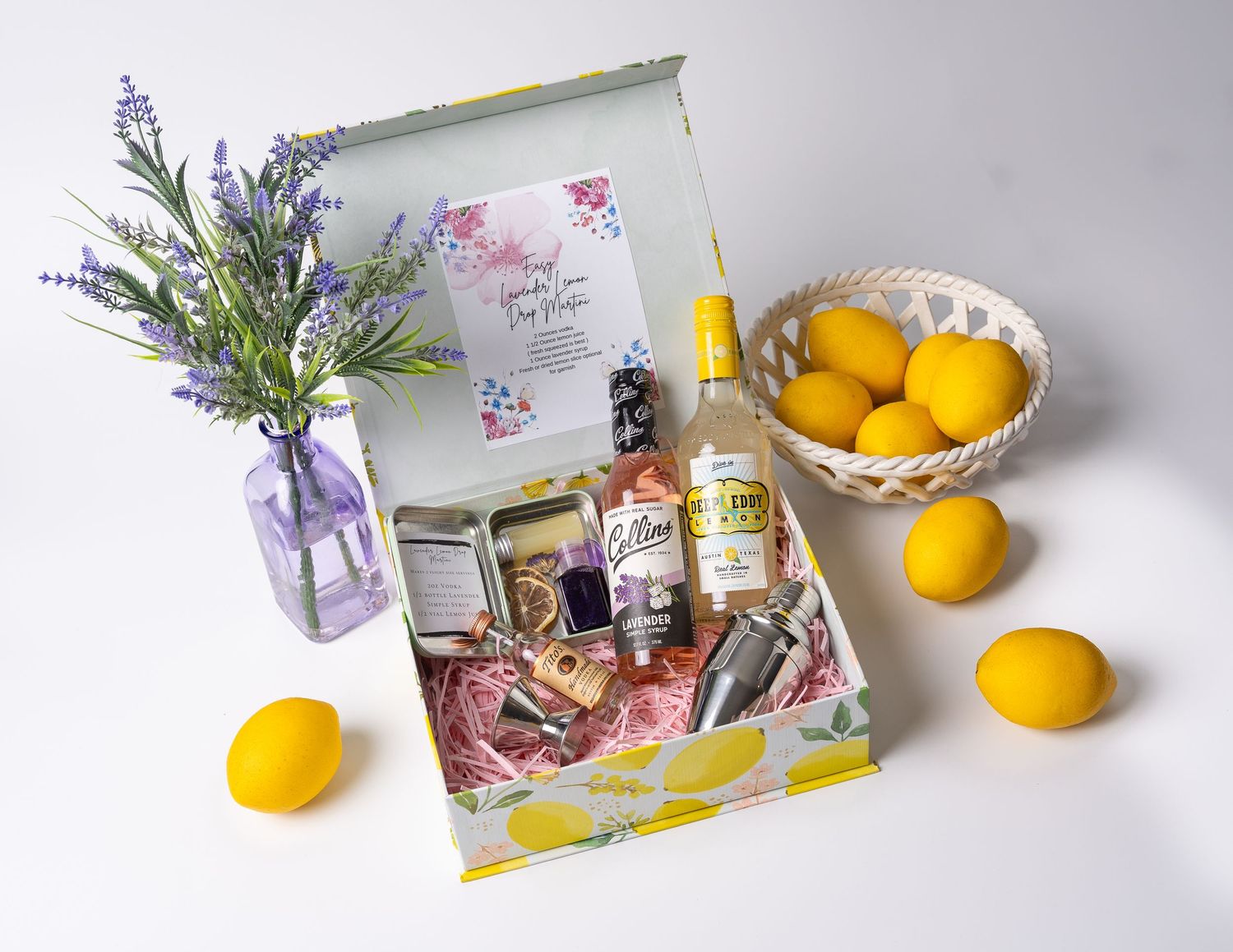 Gift box arrangement with lemons, lavender sprigs, and cocktail ingredients on white surface.