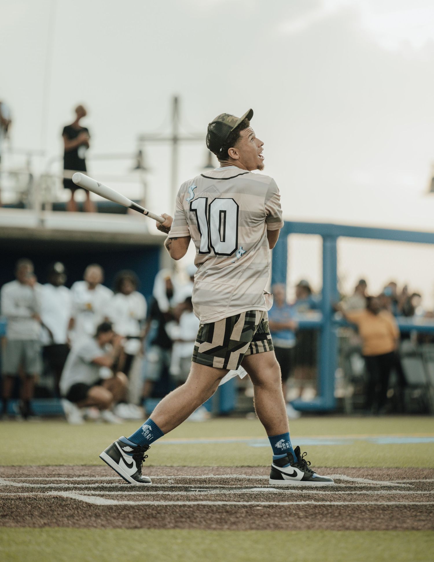 Young baseball player in jersey number 10 swings bat during game with crowd and blue dugout in background.