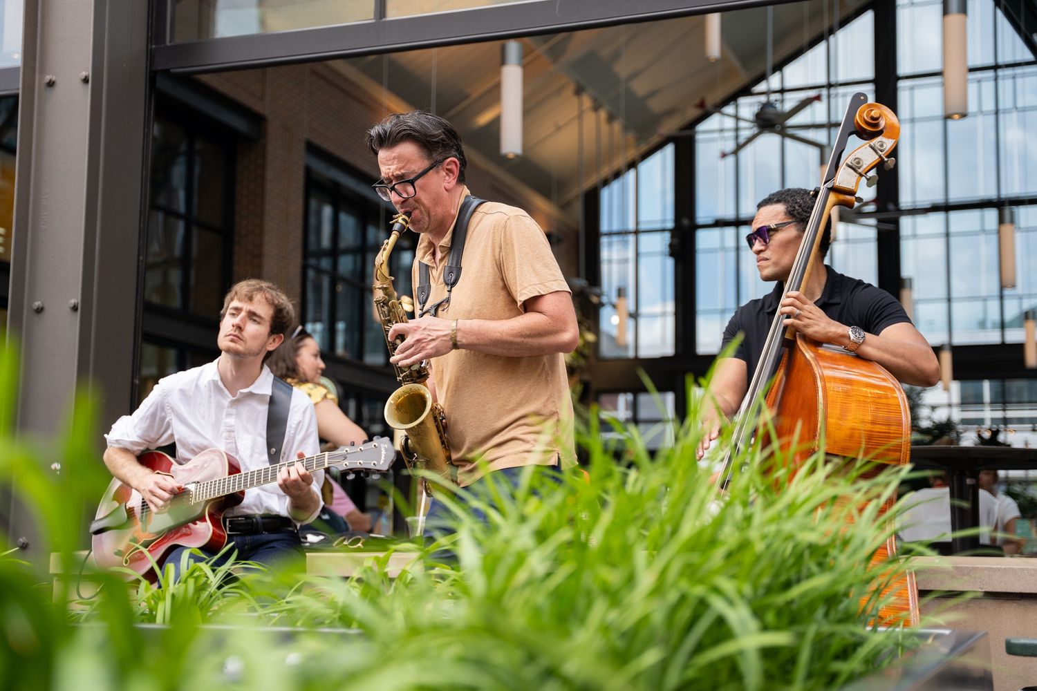Jazz musicians perform outdoors with guitar, saxophone and upright bass in front of a glass building facade.