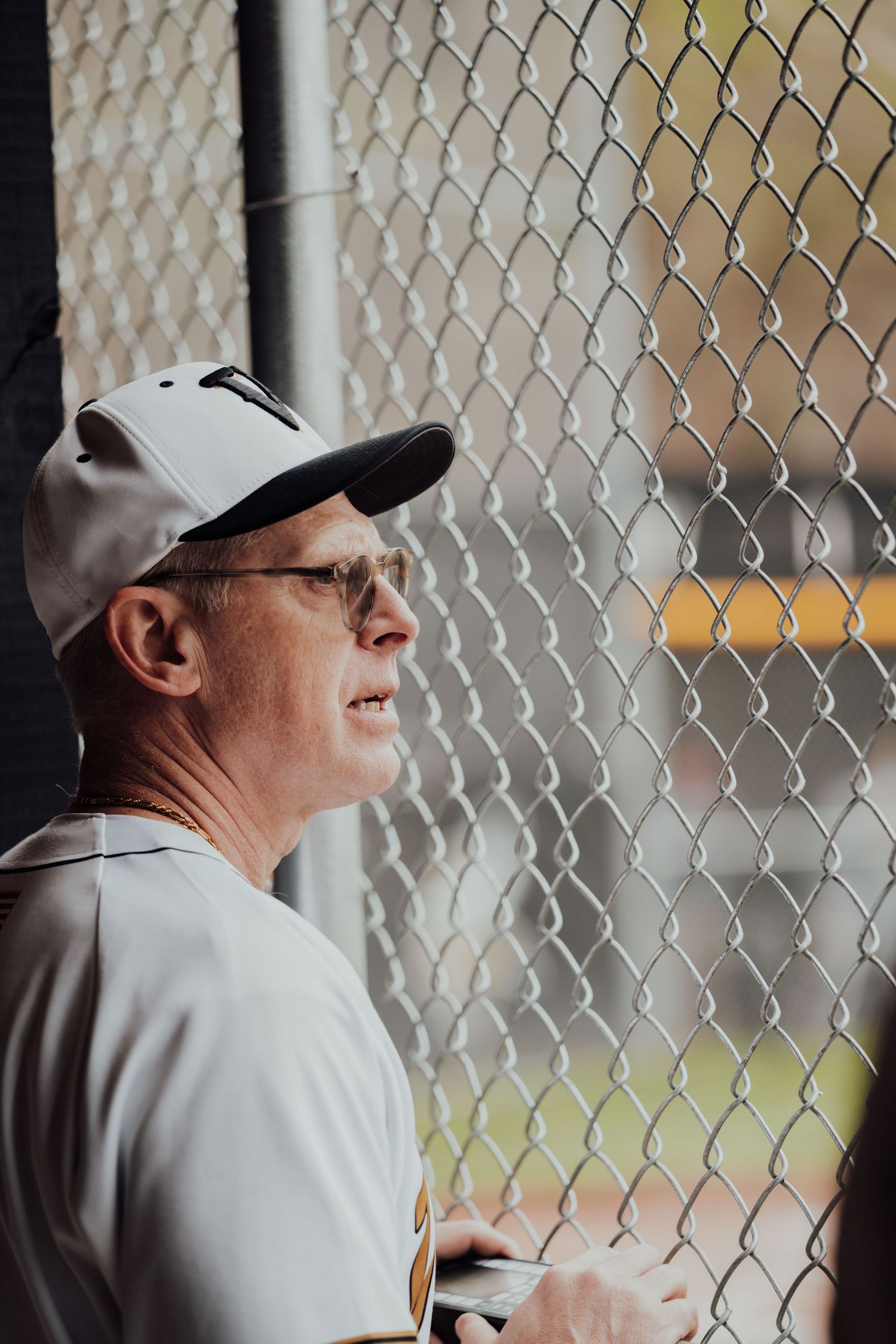 Baseball players watch game through chain link fence of dugout.