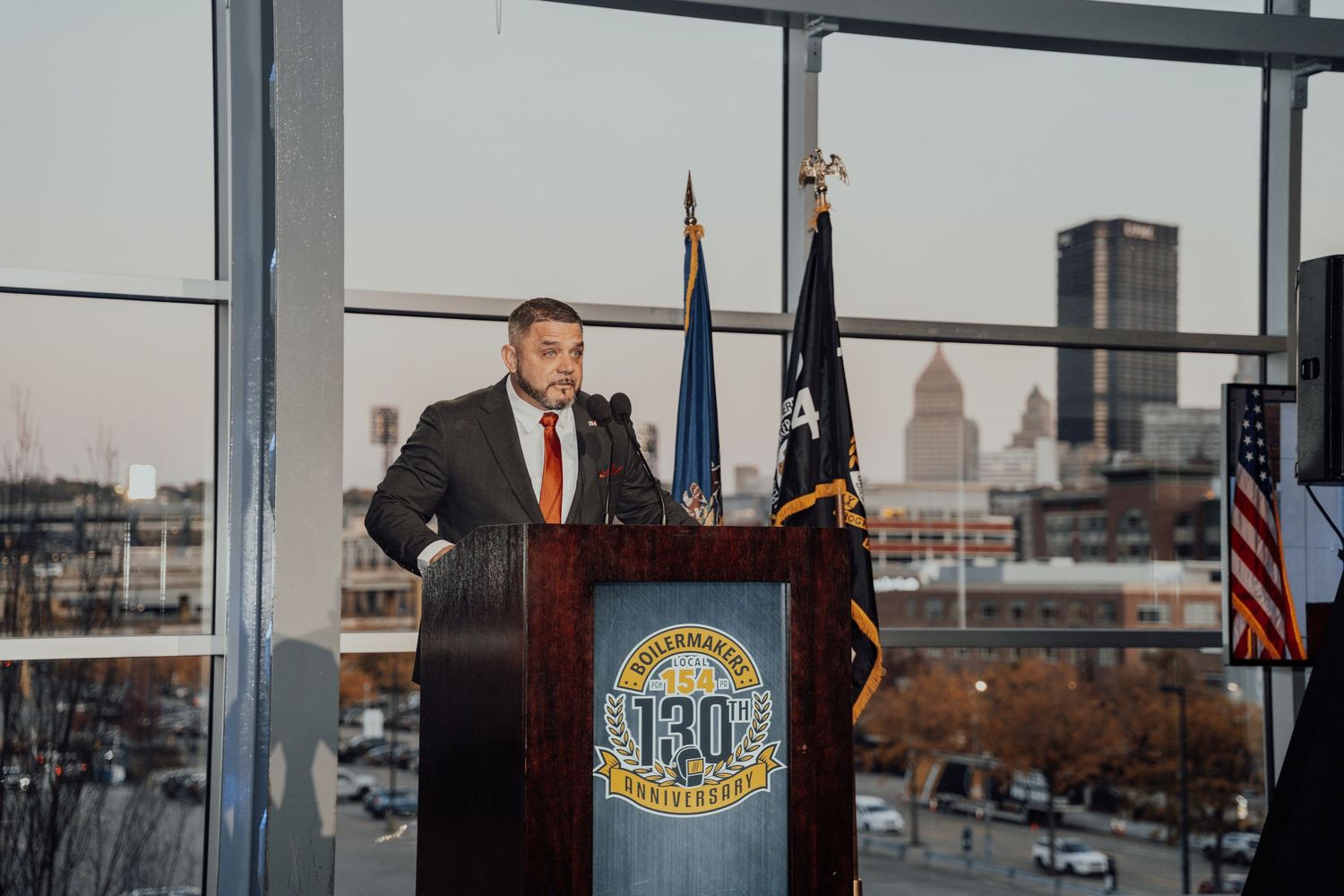 A speaker at a podium with official flags and seals during a formal ceremony with city views in the background.