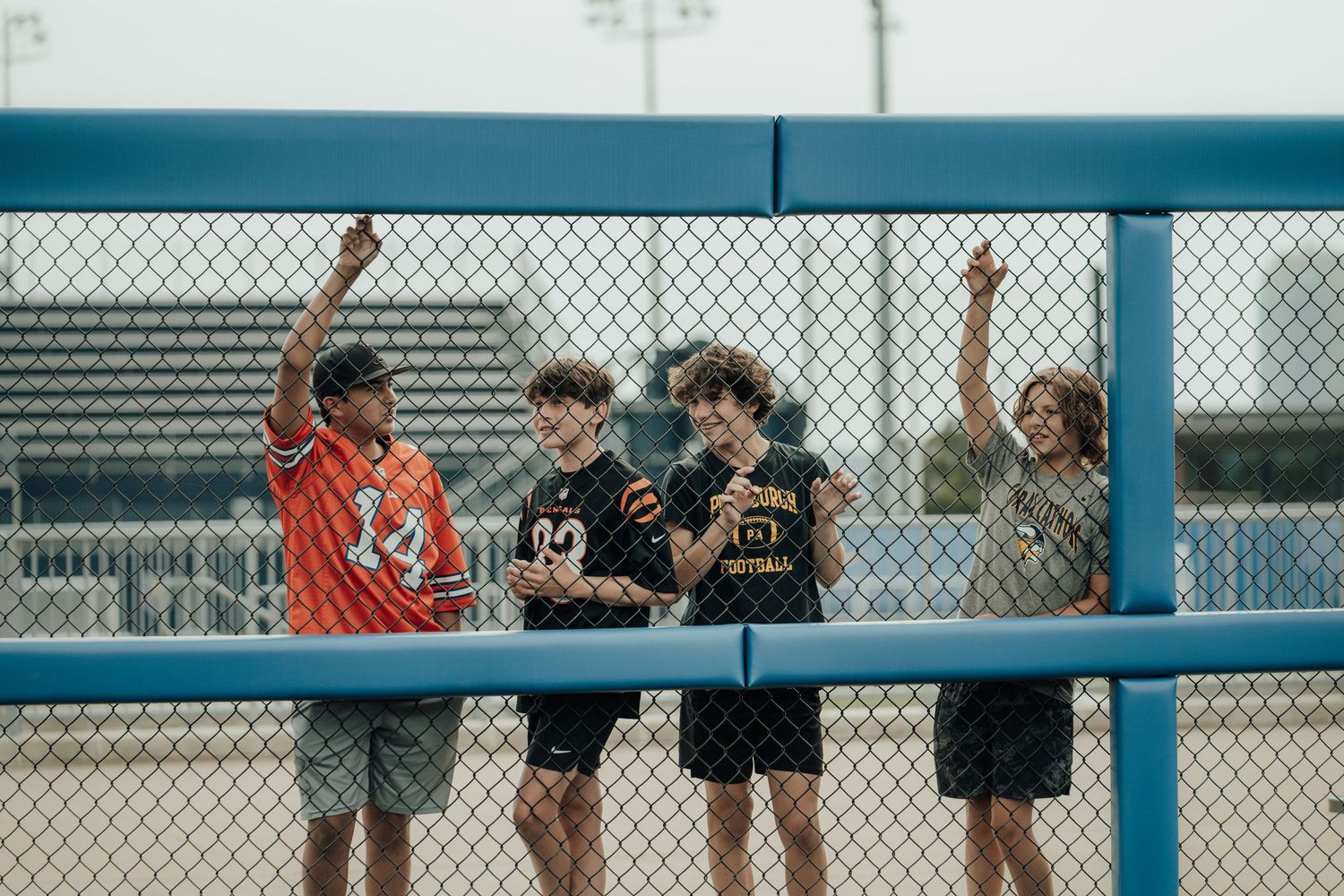 Youth baseball players and spectators watch from behind a chain link fence as a game unfolds on the field.