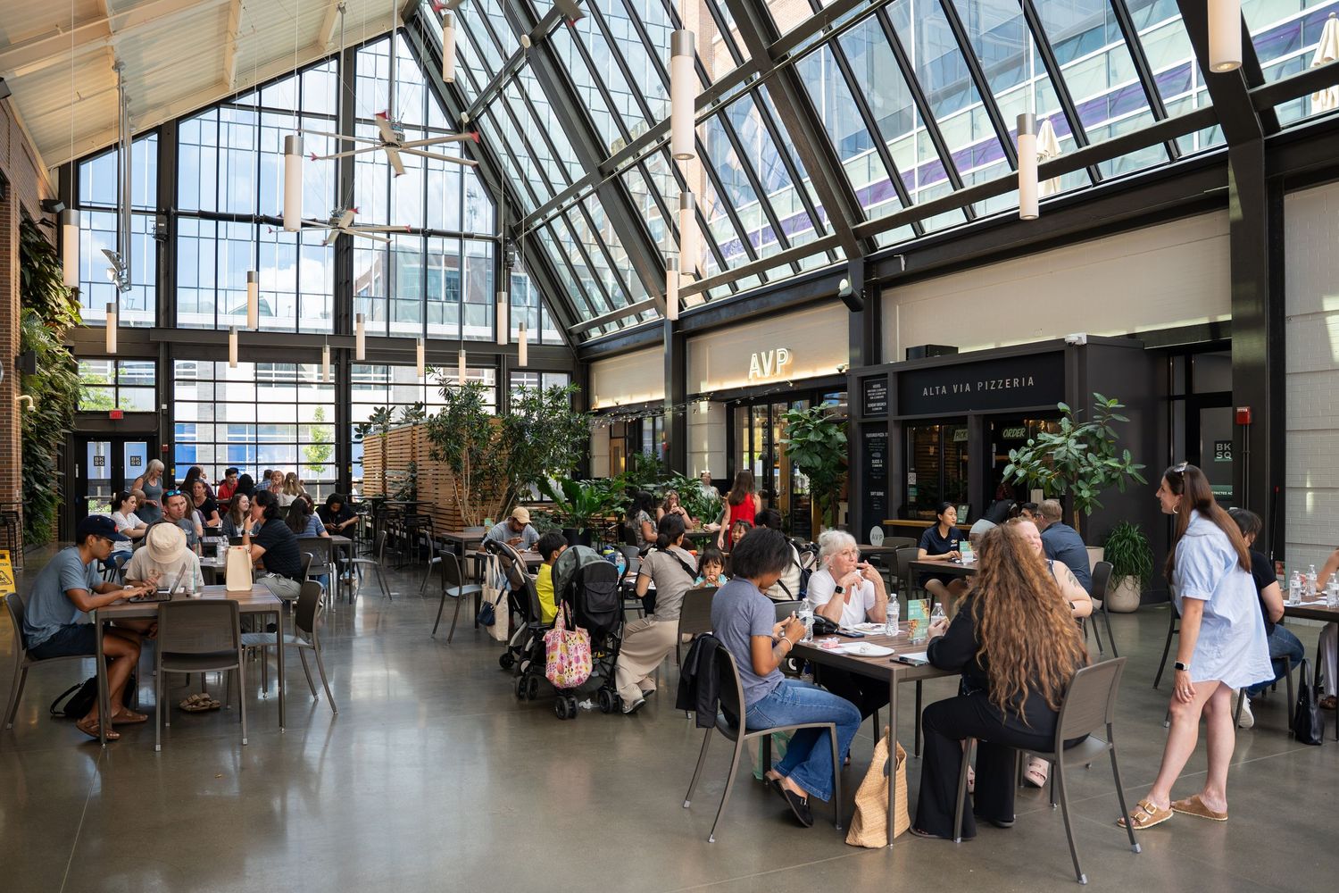 Modern atrium dining space with glass ceiling and numerous patrons seated at tables in a bright, airy setting.