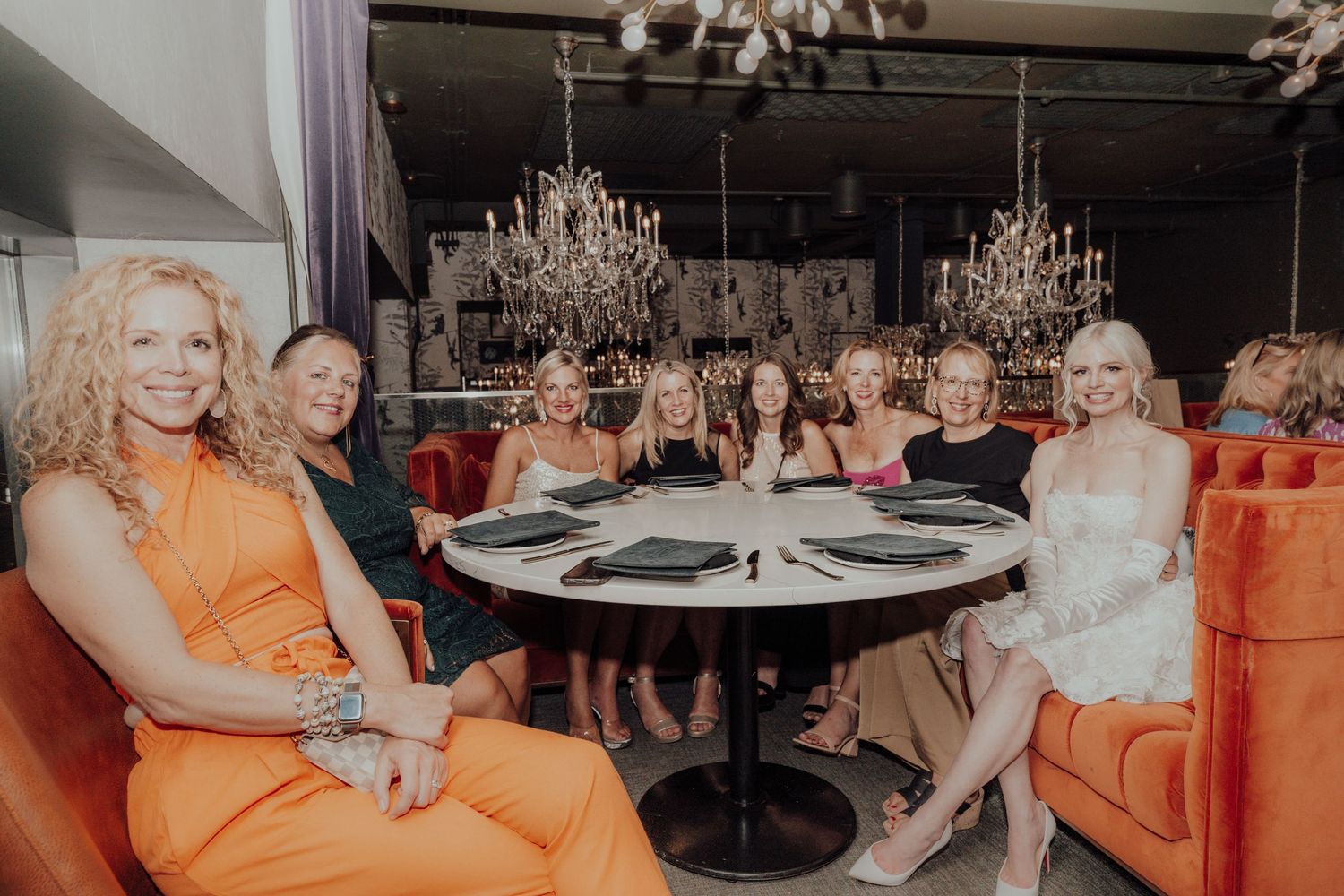A group in orange and white outfits sits around an elegant table with crystal chandeliers overhead.