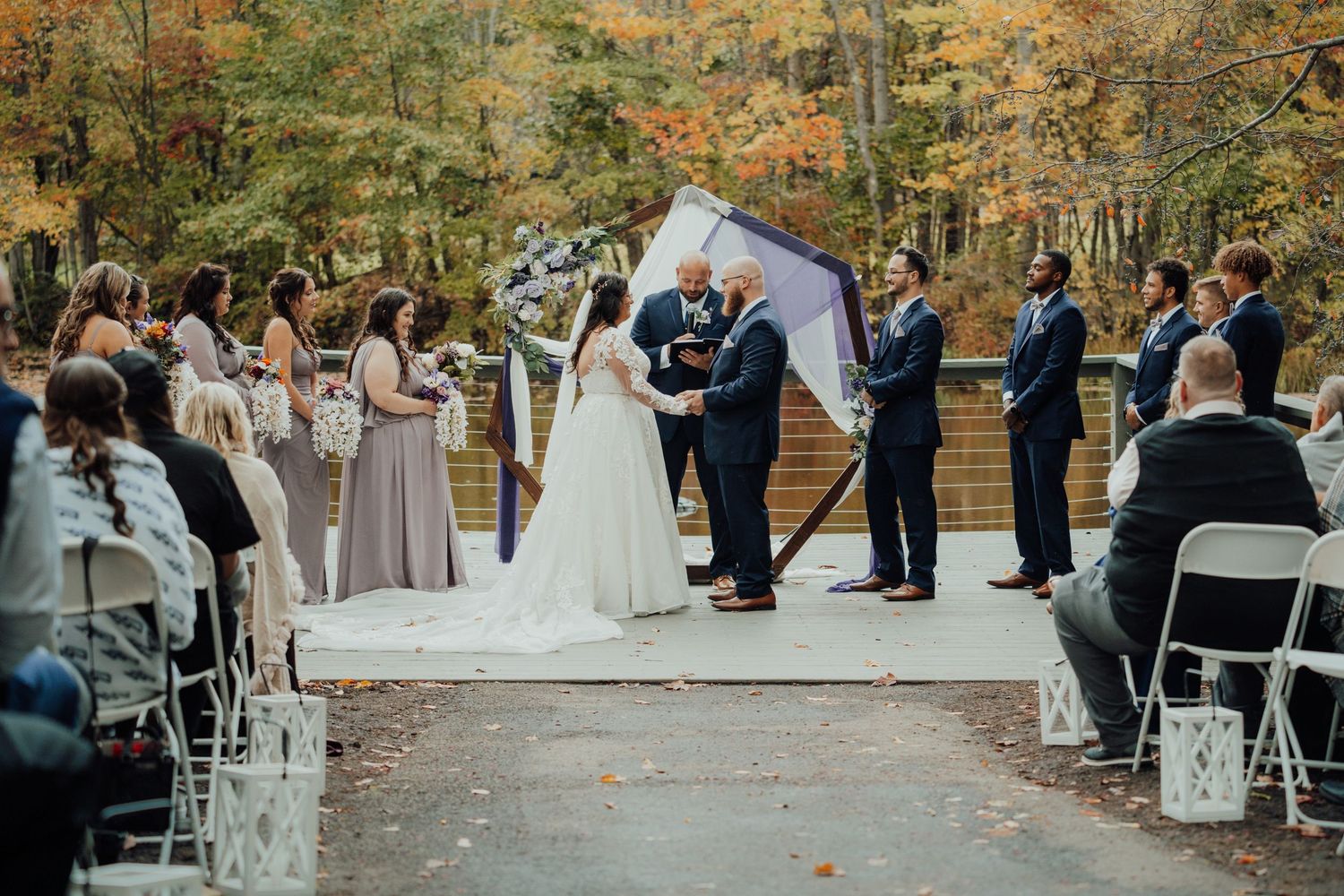 Outdoor autumn wedding ceremony with white chairs and fall foliage in the background.