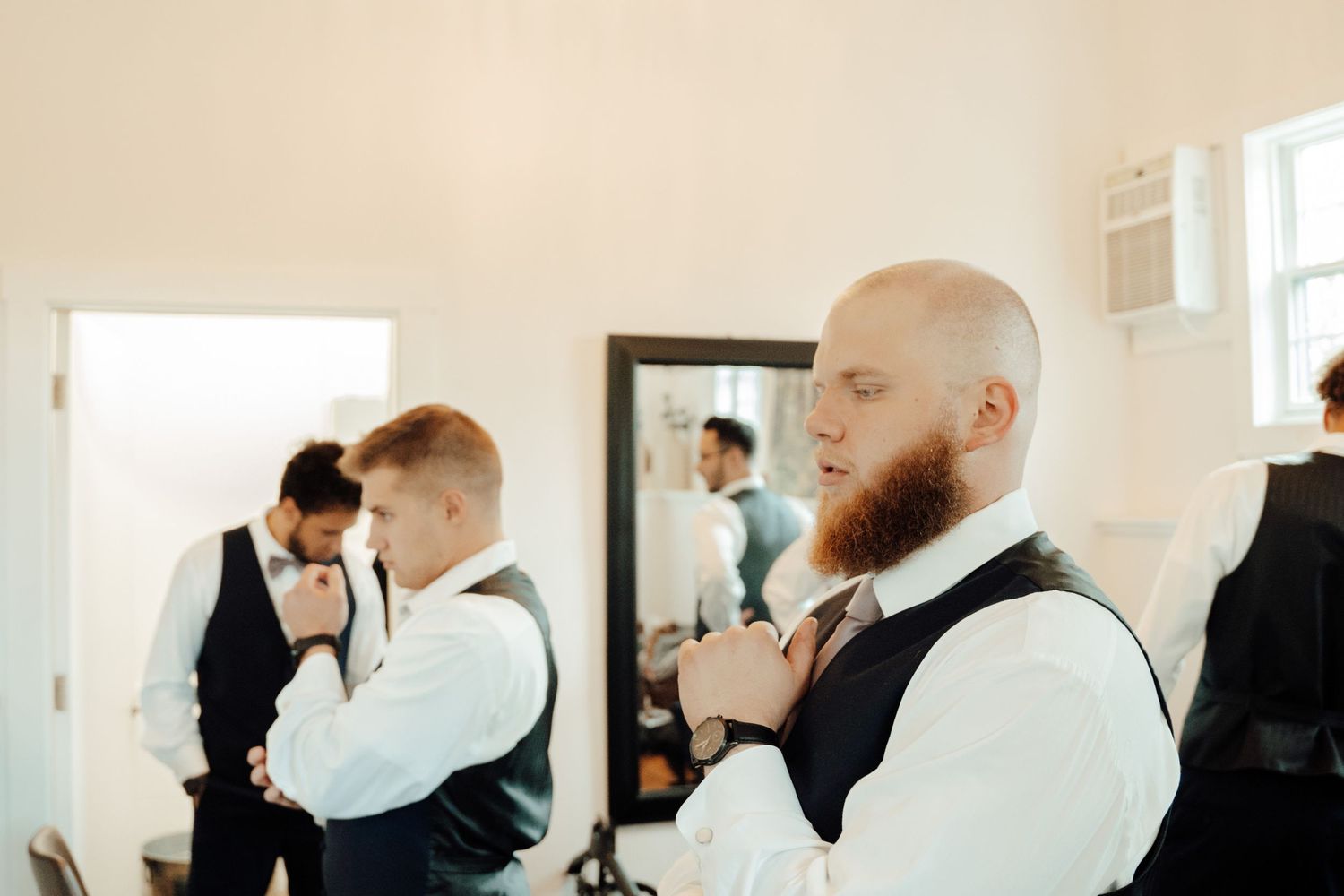 Groomsmen in vests prepare for wedding while one adjusts his tie in mirror.