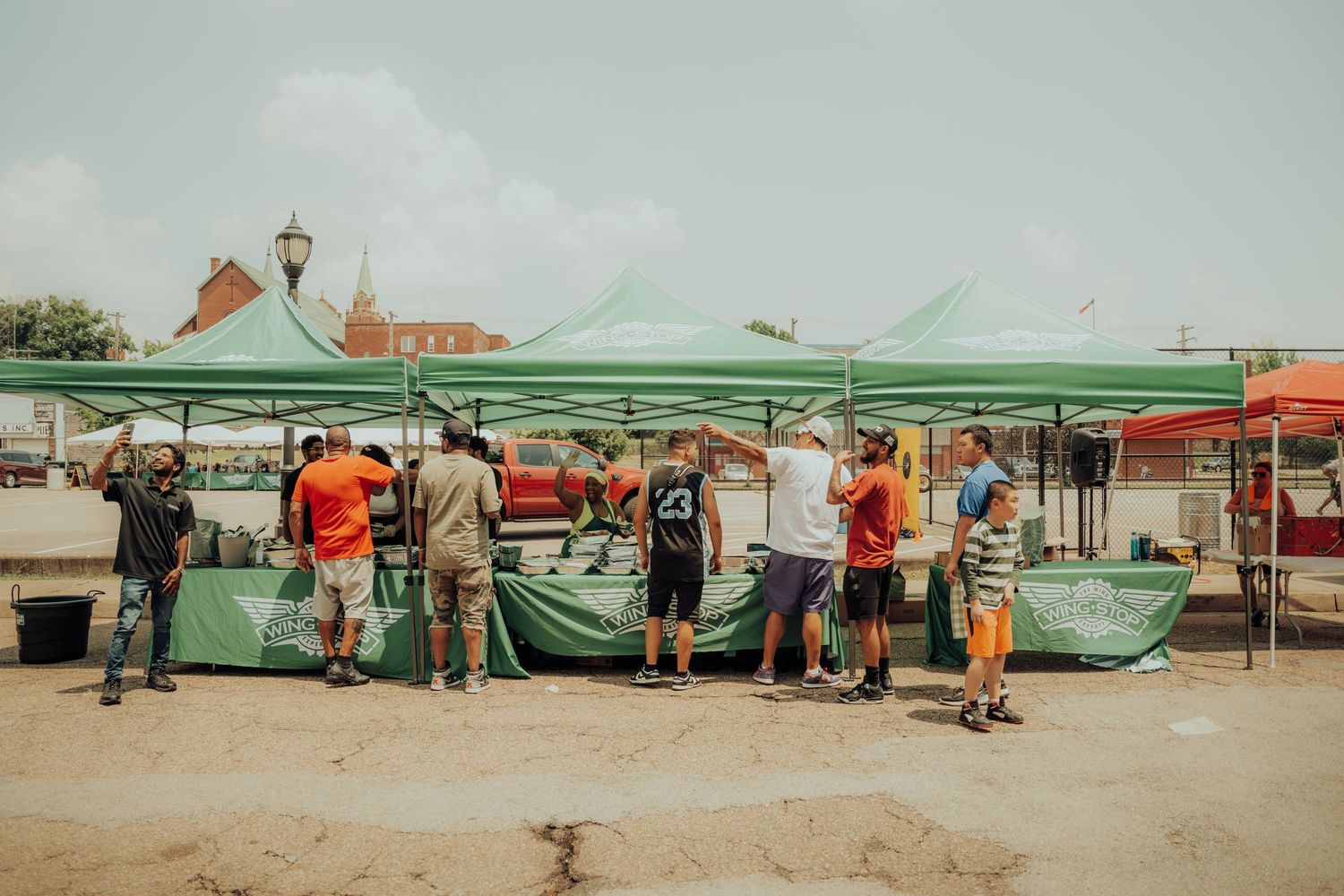 People gather under green canopy tents at an outdoor market or vendor area on a sunny day.