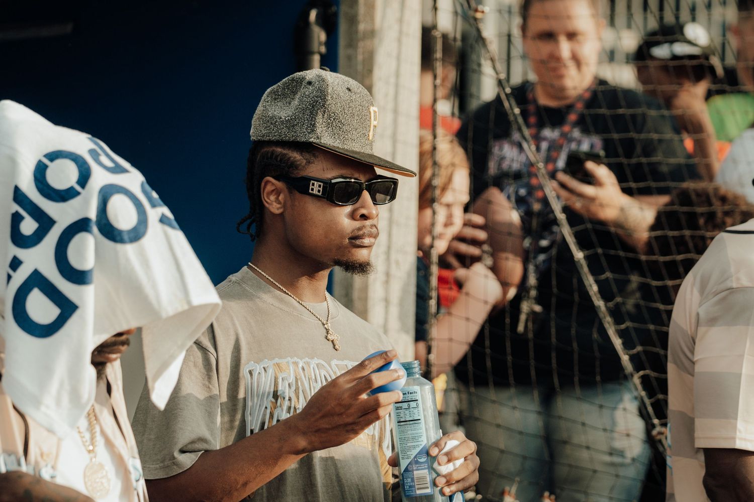 Someone in sunglasses and baseball cap holds a water bottle while standing near a fence at an outdoor event.