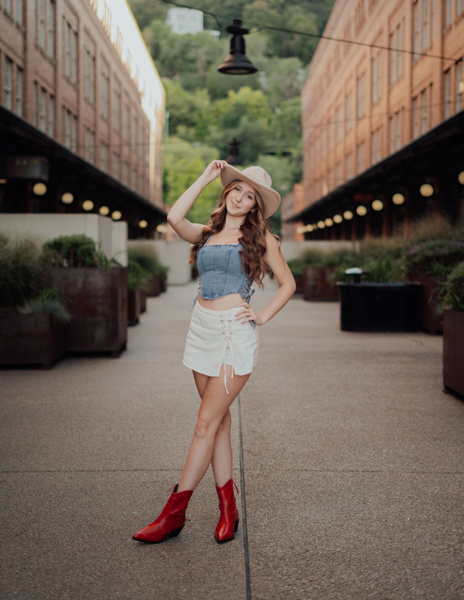 A fashionable young woman in a crop top and white skirt poses on an urban walkway wearing a sun hat and red boots.