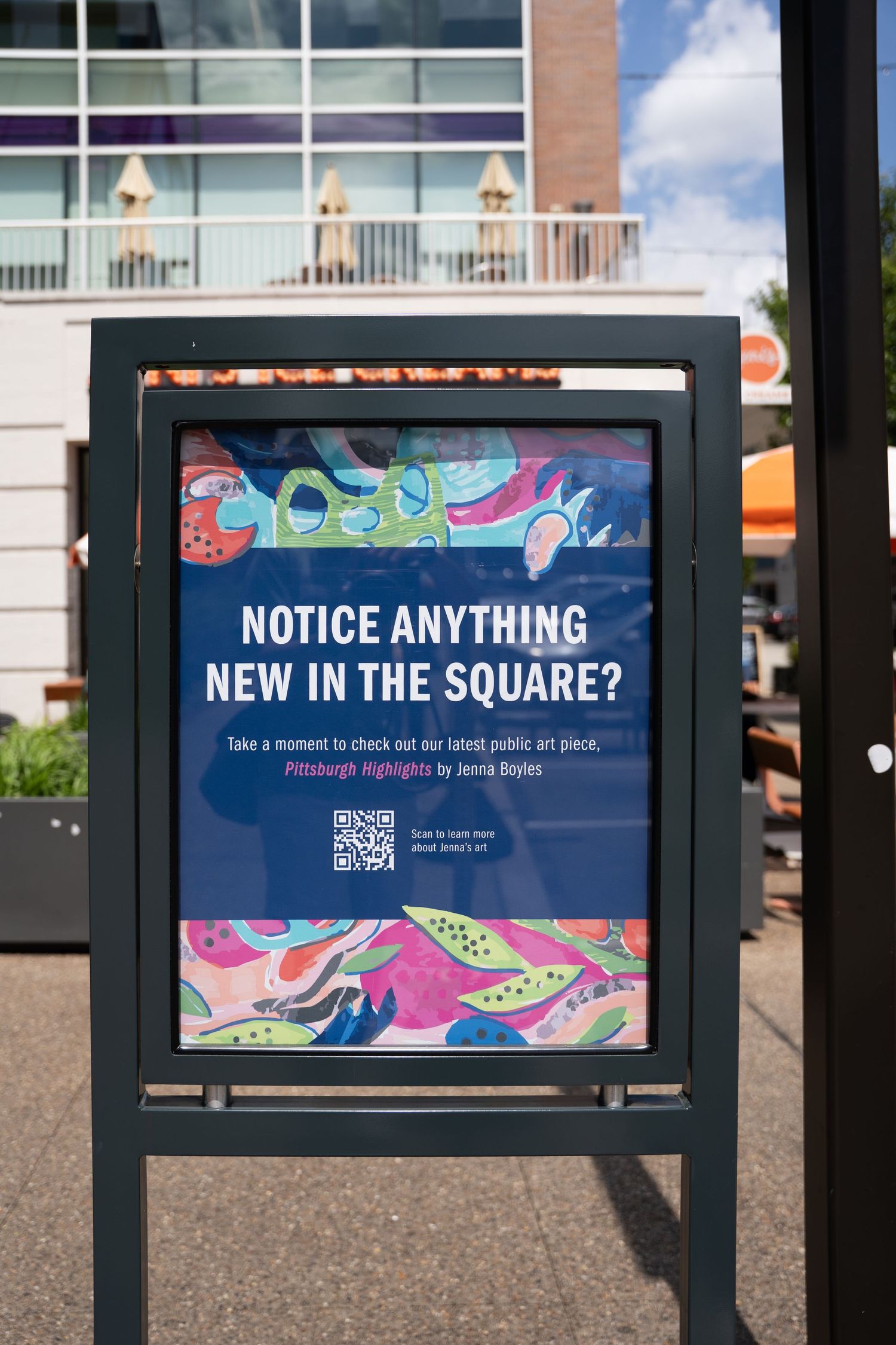 A colorful public notice sign asking 'Notice anything new in the square?' with decorative artwork and a QR code.
