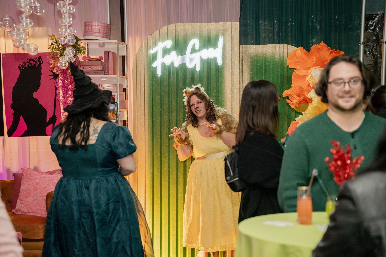 Event guests mingle around a colorful rainbow backdrop with neon signage and tropical decorations at an indoor party.
