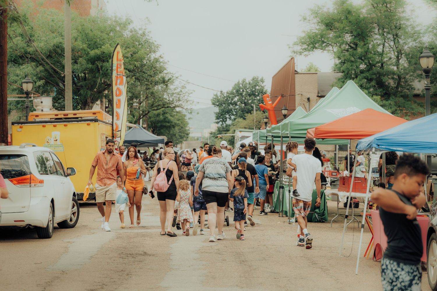 A busy summer street festival with colorful vendor tents and crowds walking between food stalls and market booths.