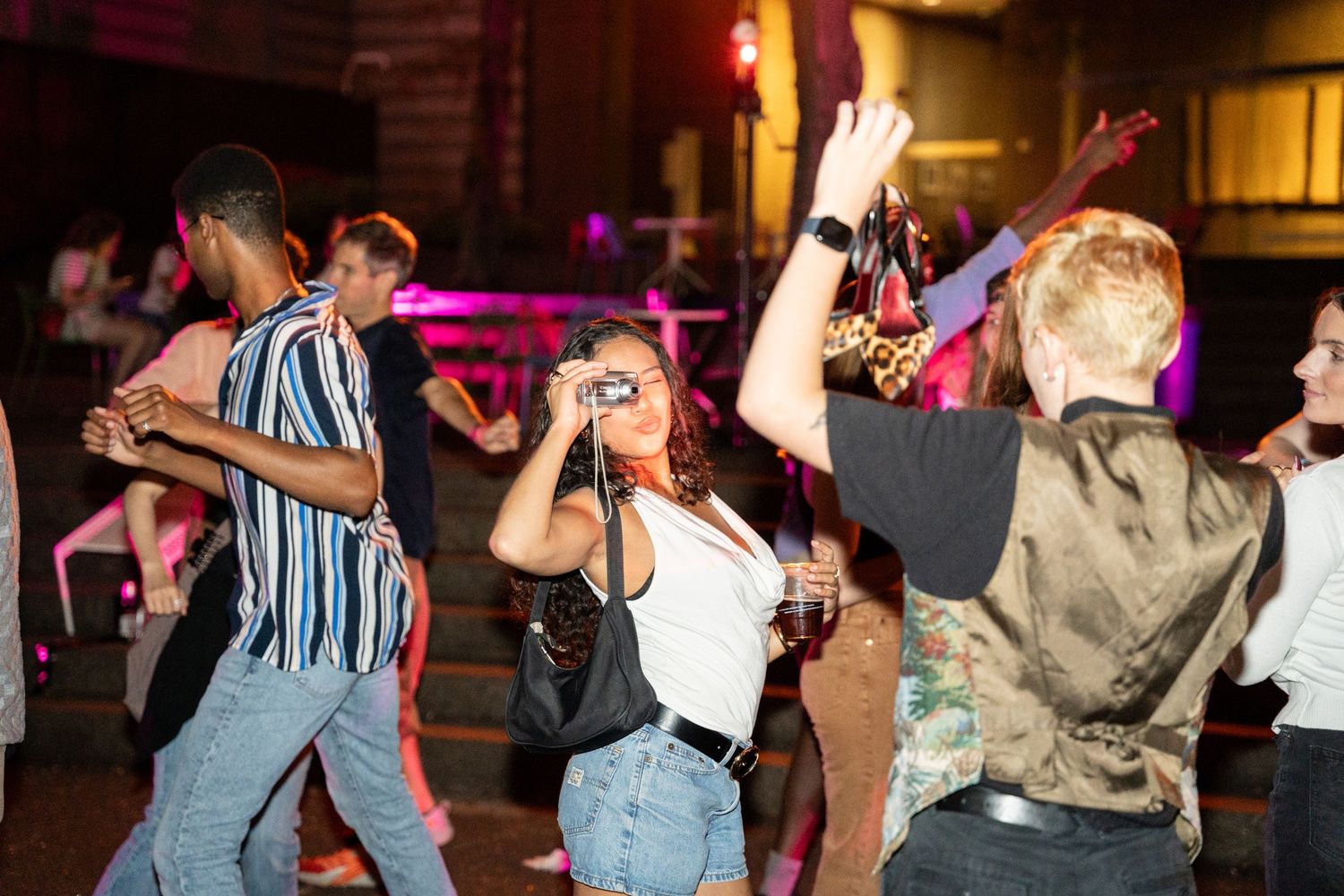 People dancing and socializing at a nighttime party with pink and purple lighting.