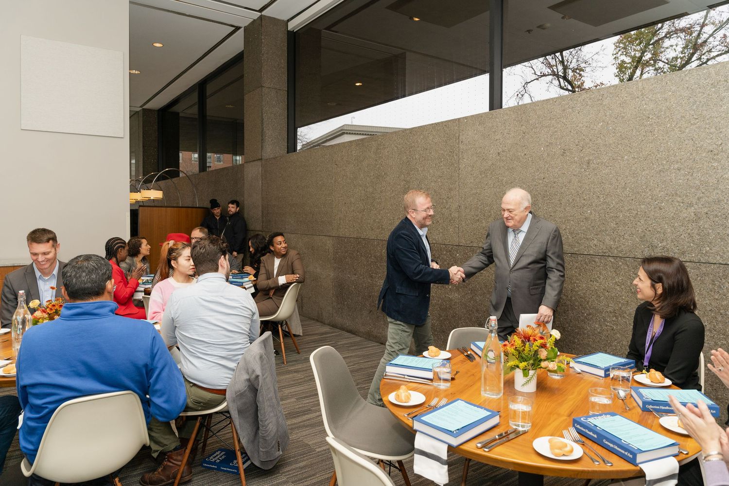 Business meeting attendees gather around tables with orange centerpieces in a modern dining space.