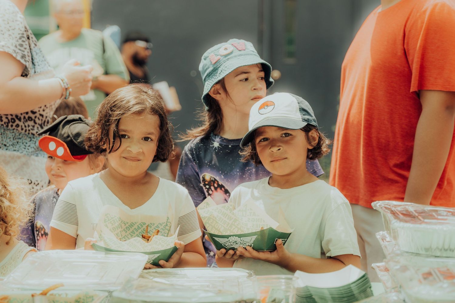 Children gather around a table participating in an outdoor arts and crafts activity on a sunny summer day.