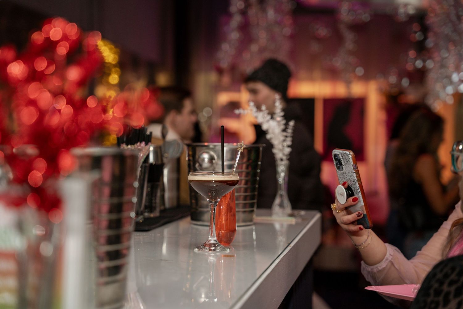Cocktail glass sits on bar counter while someone takes photo with phone in dimly lit venue with red bokeh lights.