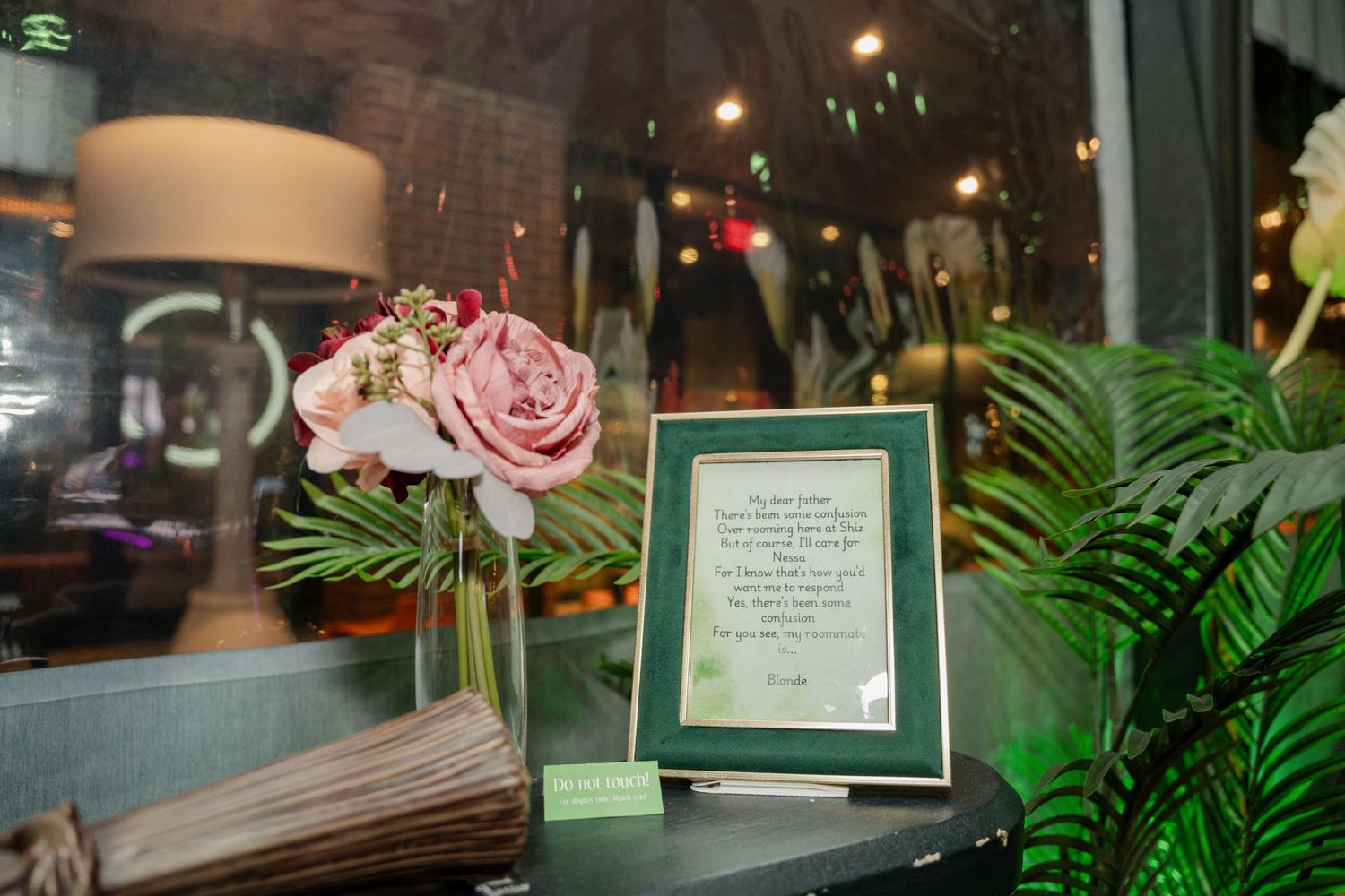 Elegant table setting with pink roses in a vase, framed document, and tropical plants in a dimly lit interior.