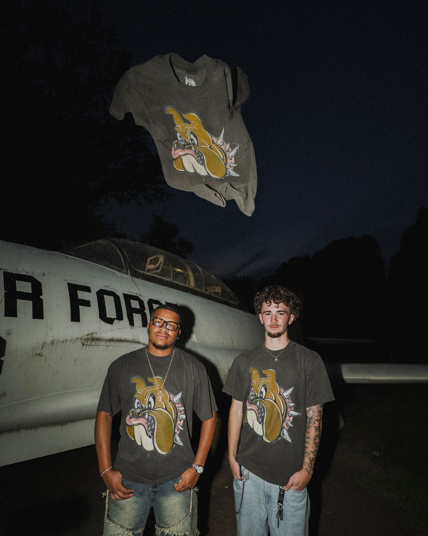 Two people posing in matching bulldog graphic t-shirts in front of a military aircraft at night.