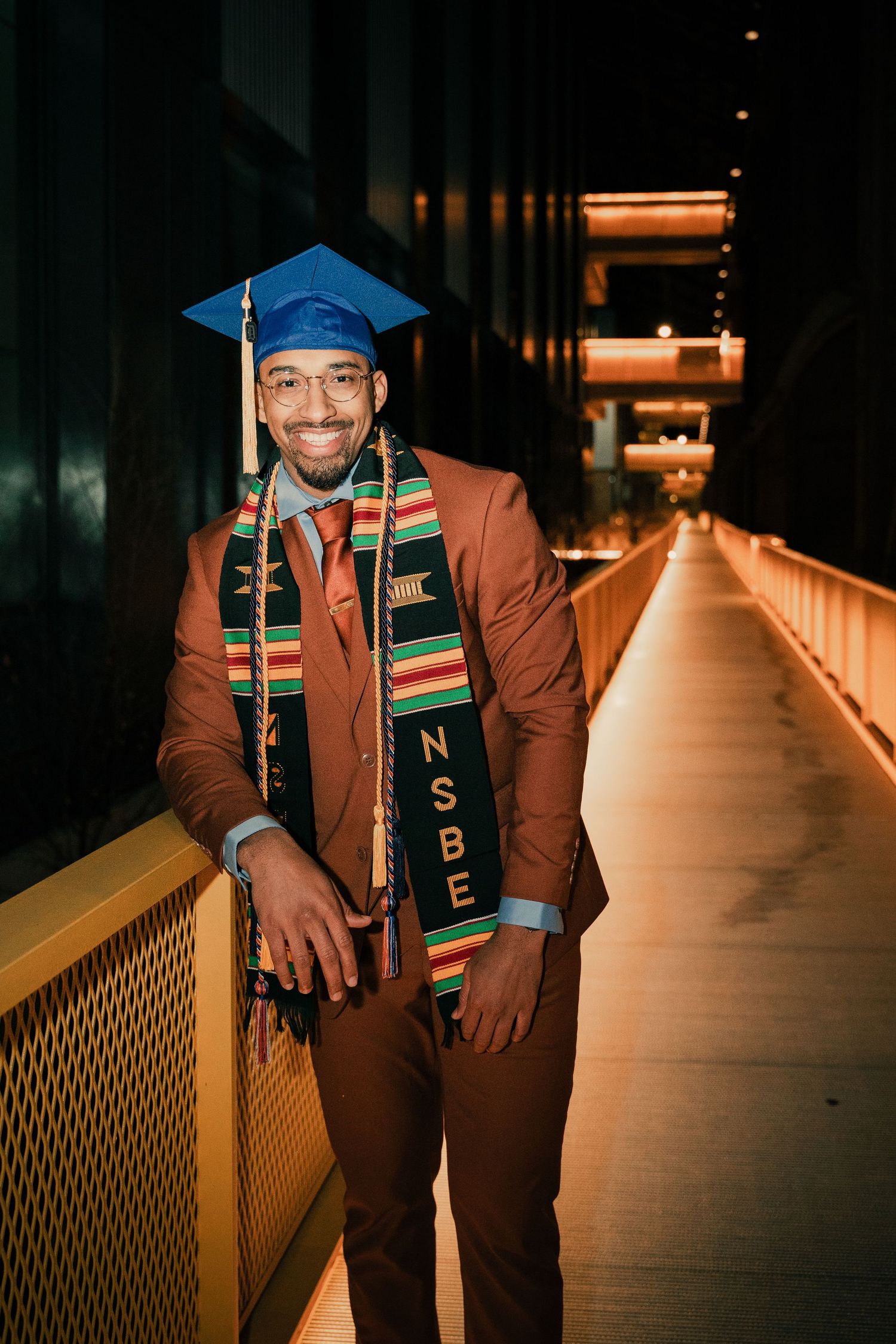 Graduate in blue cap and brown suit poses with traditional kente stole and honors decorations in illuminated hallway.