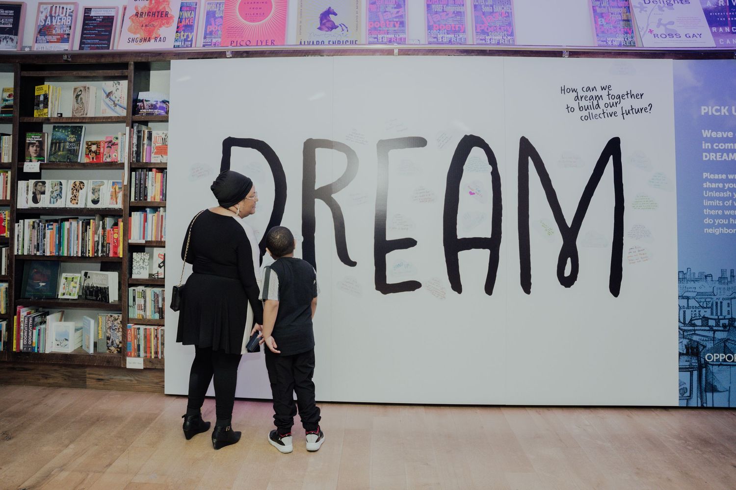 Row of colorful posters and artwork displayed on a wall with people viewing the exhibition.