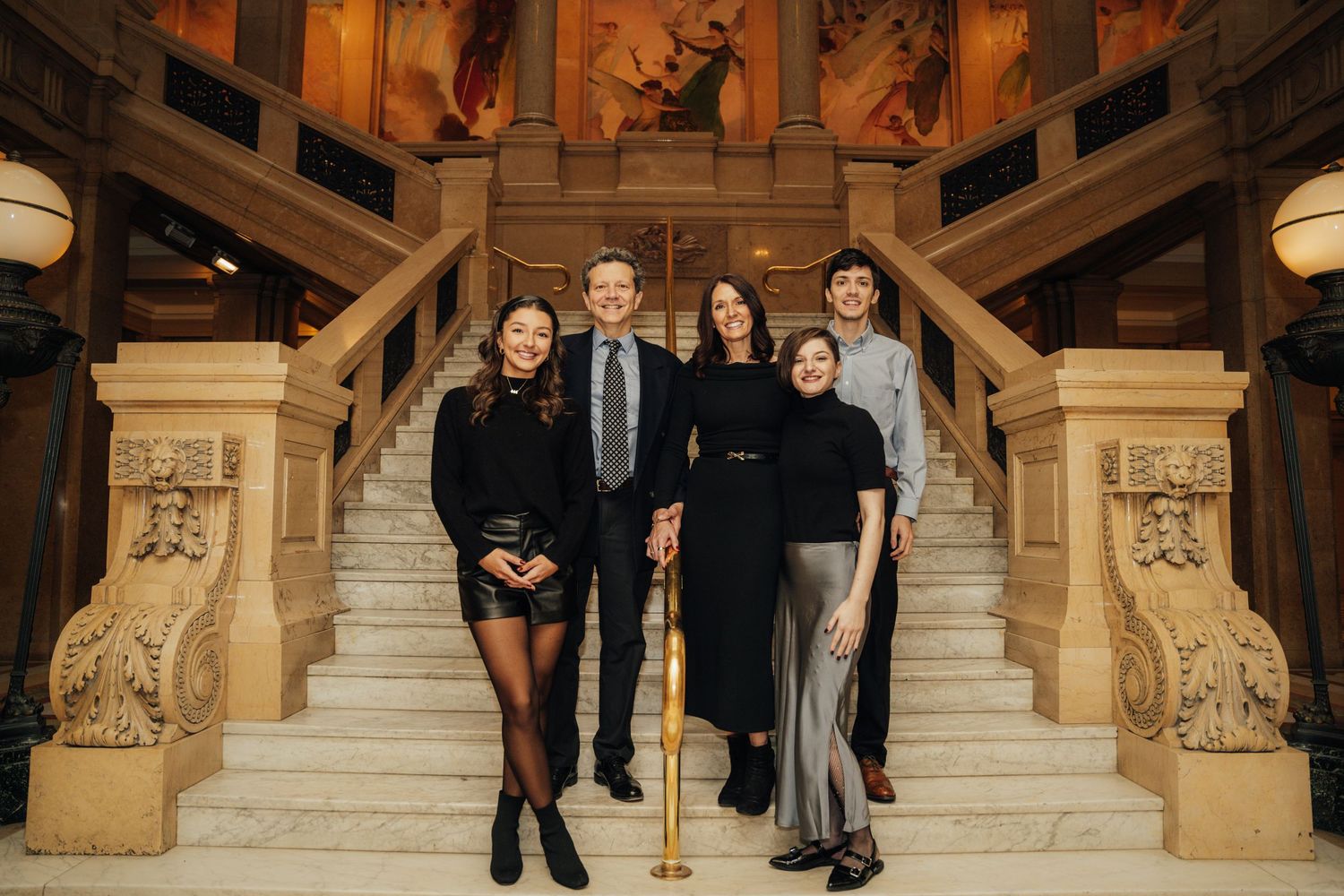 A group of formally dressed people stand on an elegant stone staircase in what appears to be a grand historic building.