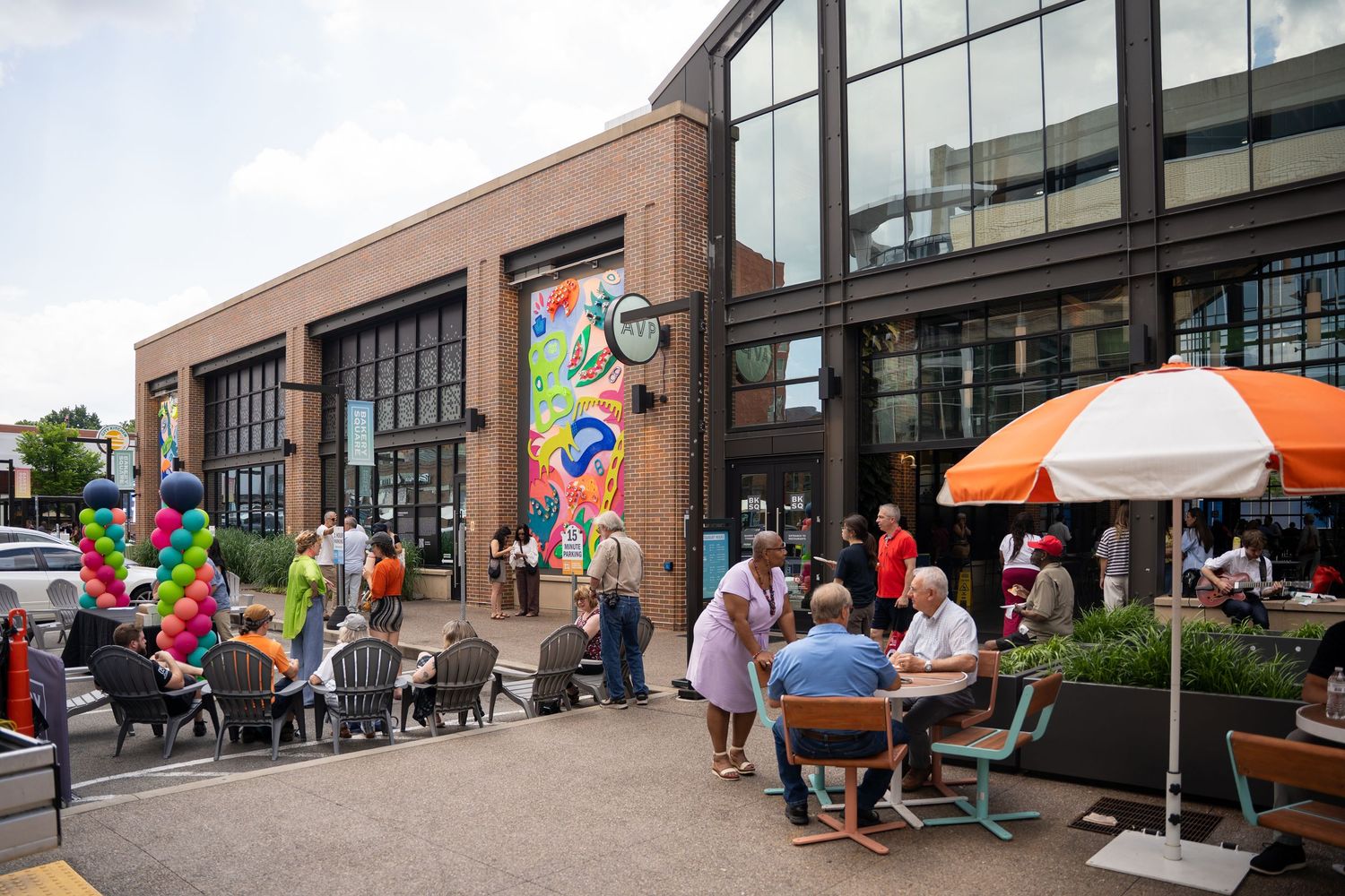 Panoramic view of a brick building exterior decorated with colorful balloon arches and people gathering for an outdoor event.