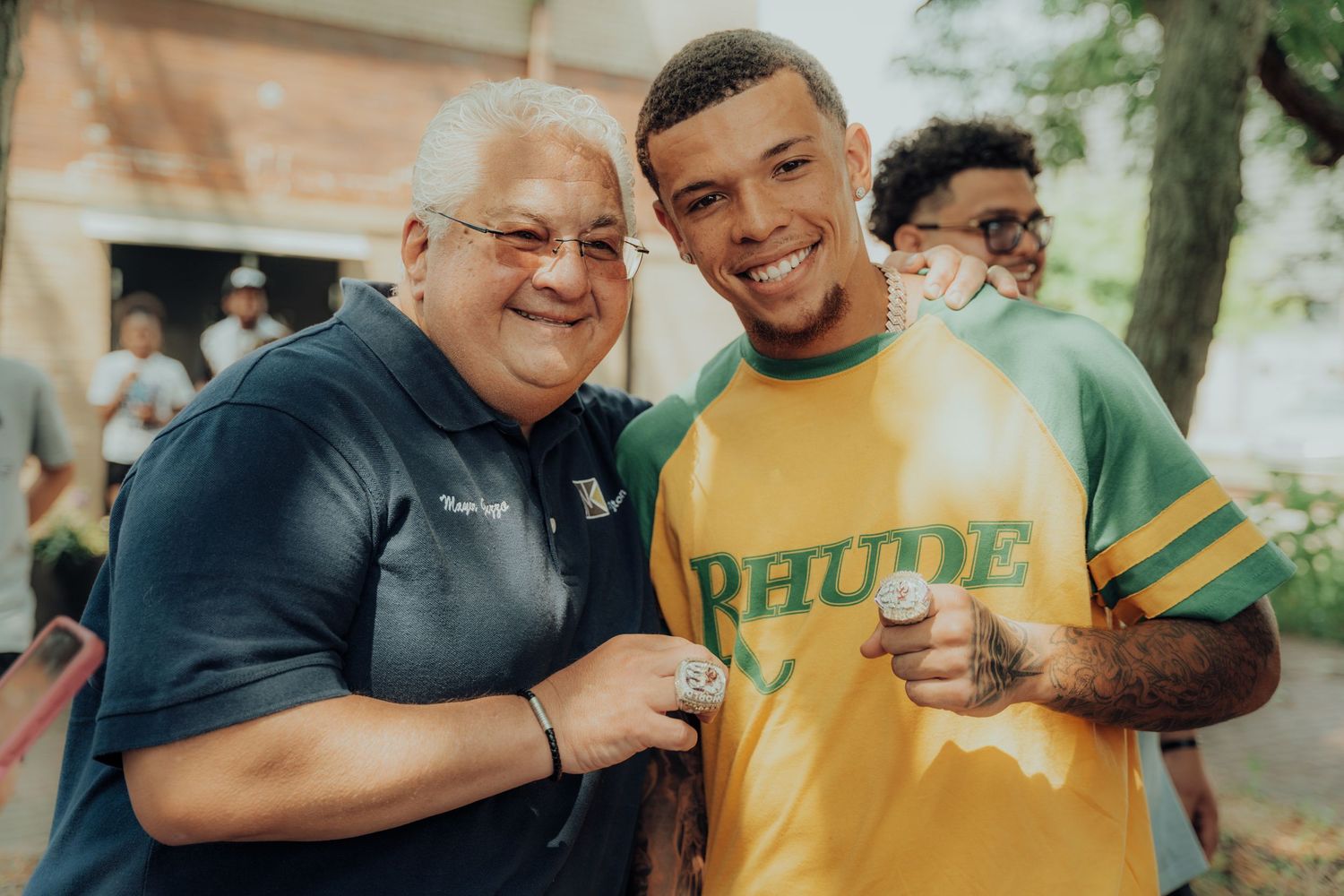 Two people smile together at an outdoor event, one wearing a navy blue shirt and one in a yellow Bruins jersey.