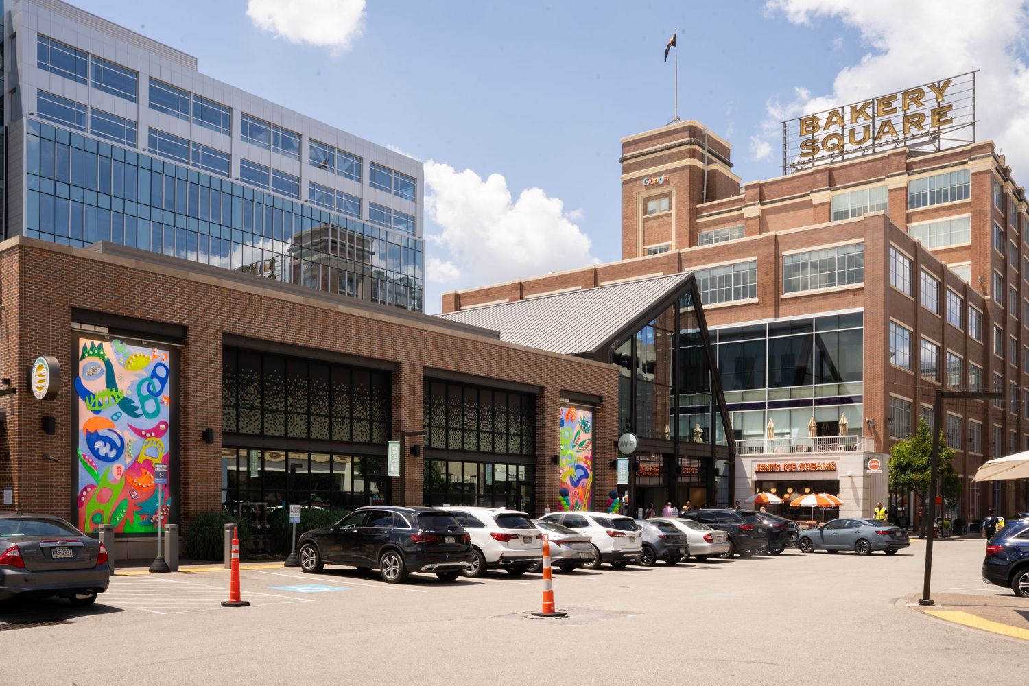 Urban shopping center exterior with retail buildings, glass facades, and parking area beneath blue skies in a downtown setting.