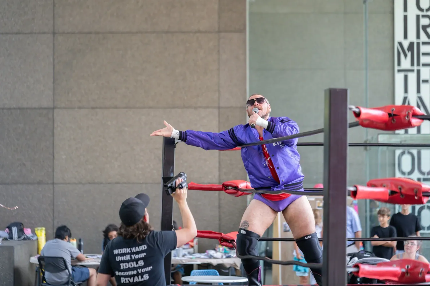 Professional wrestlers perform athletic moves in an outdoor ring during a daytime event.