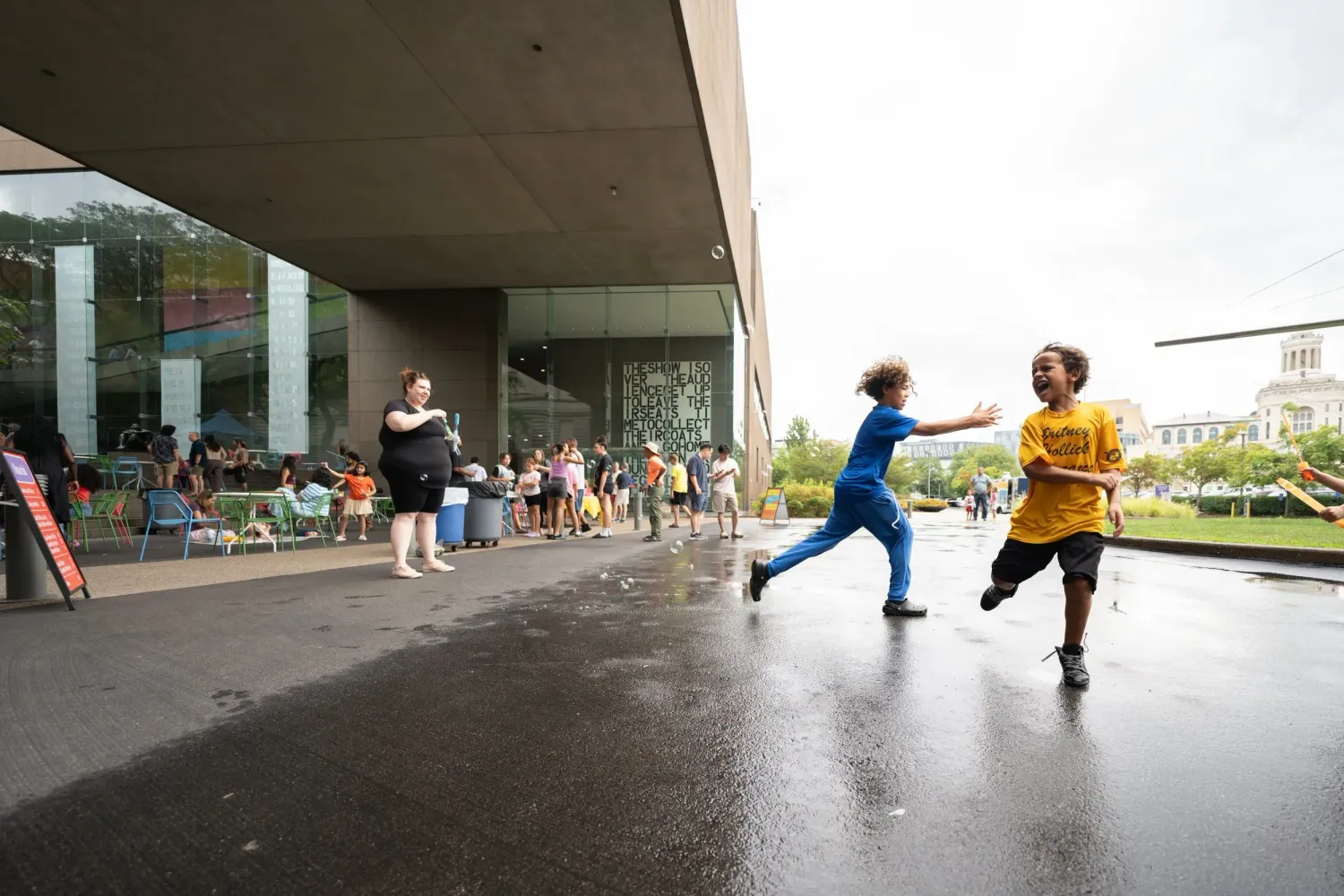 People dance joyfully under a covered walkway outside on a rainy day at a modern building entrance.