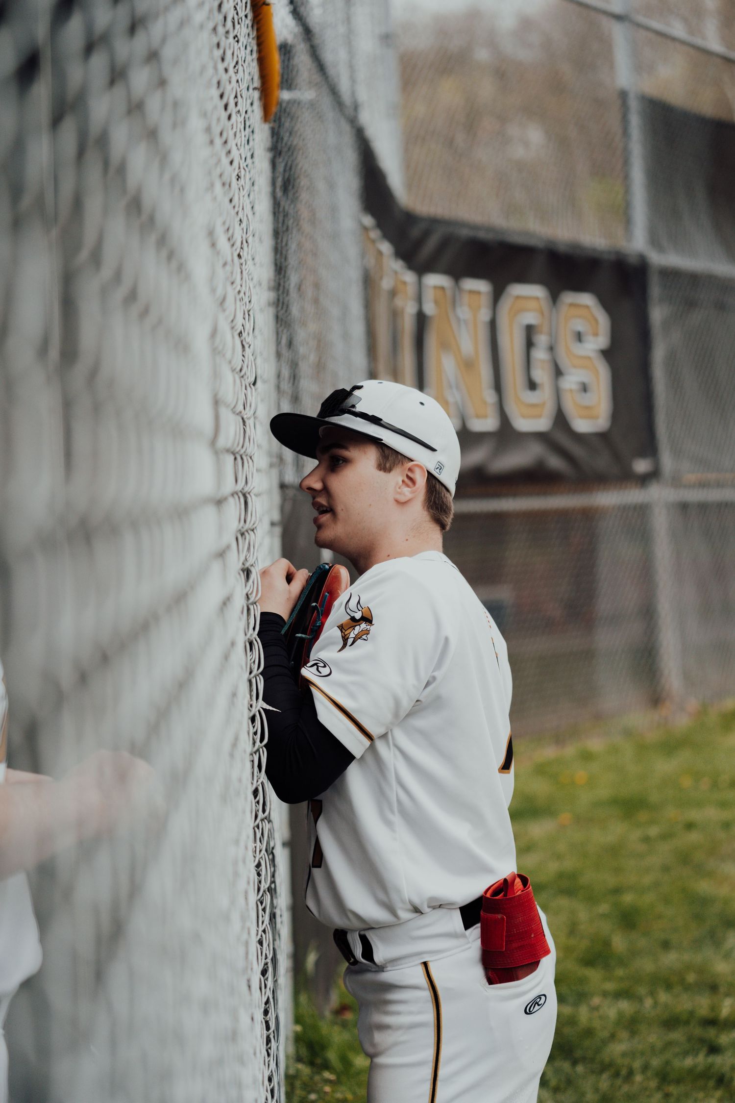 Young player in white baseball uniform peers through chain link dugout fence.