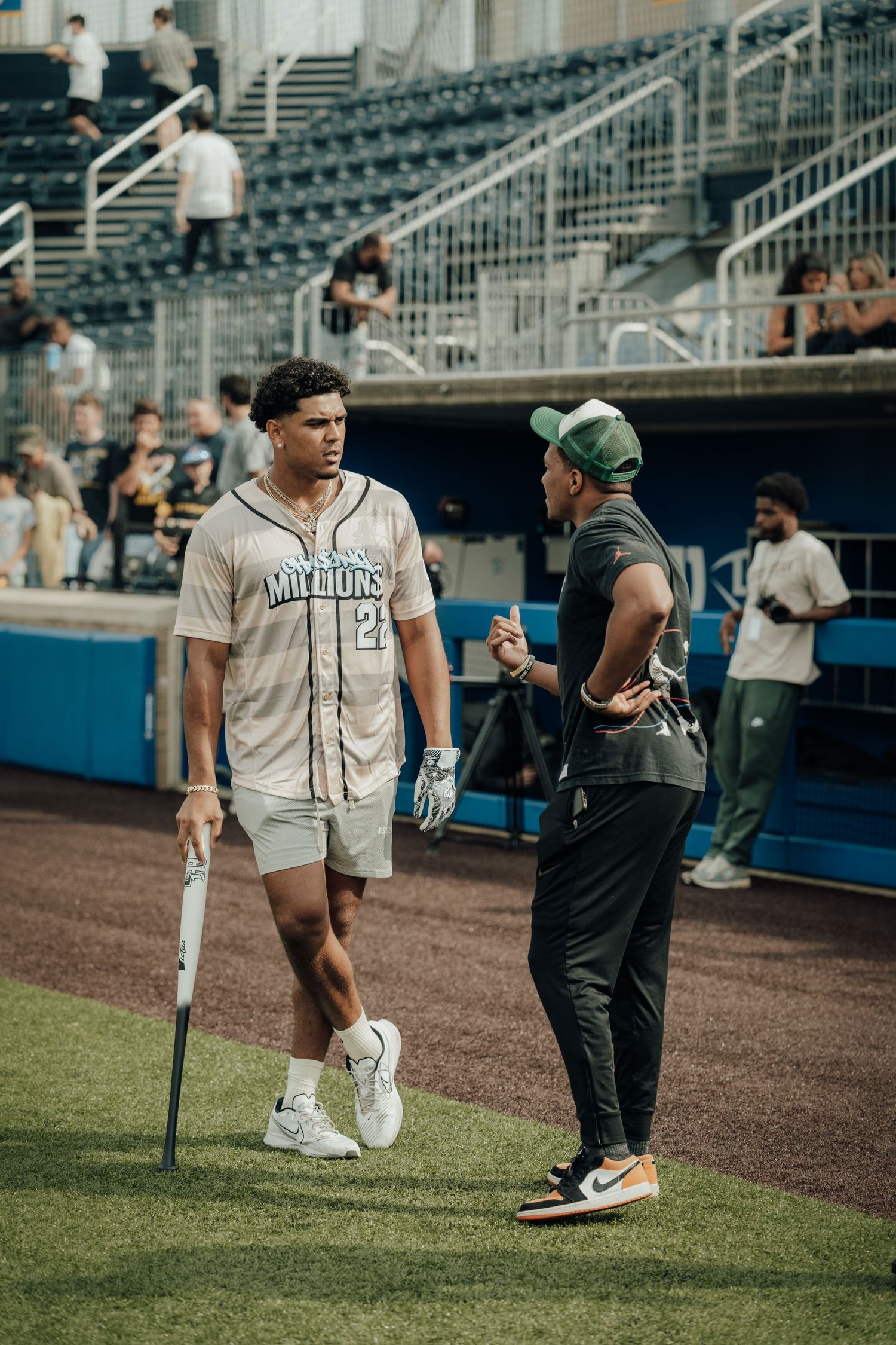 Two people chatting on a baseball field near stadium seating, one in a jersey and holding a bat and the other in dark attire.