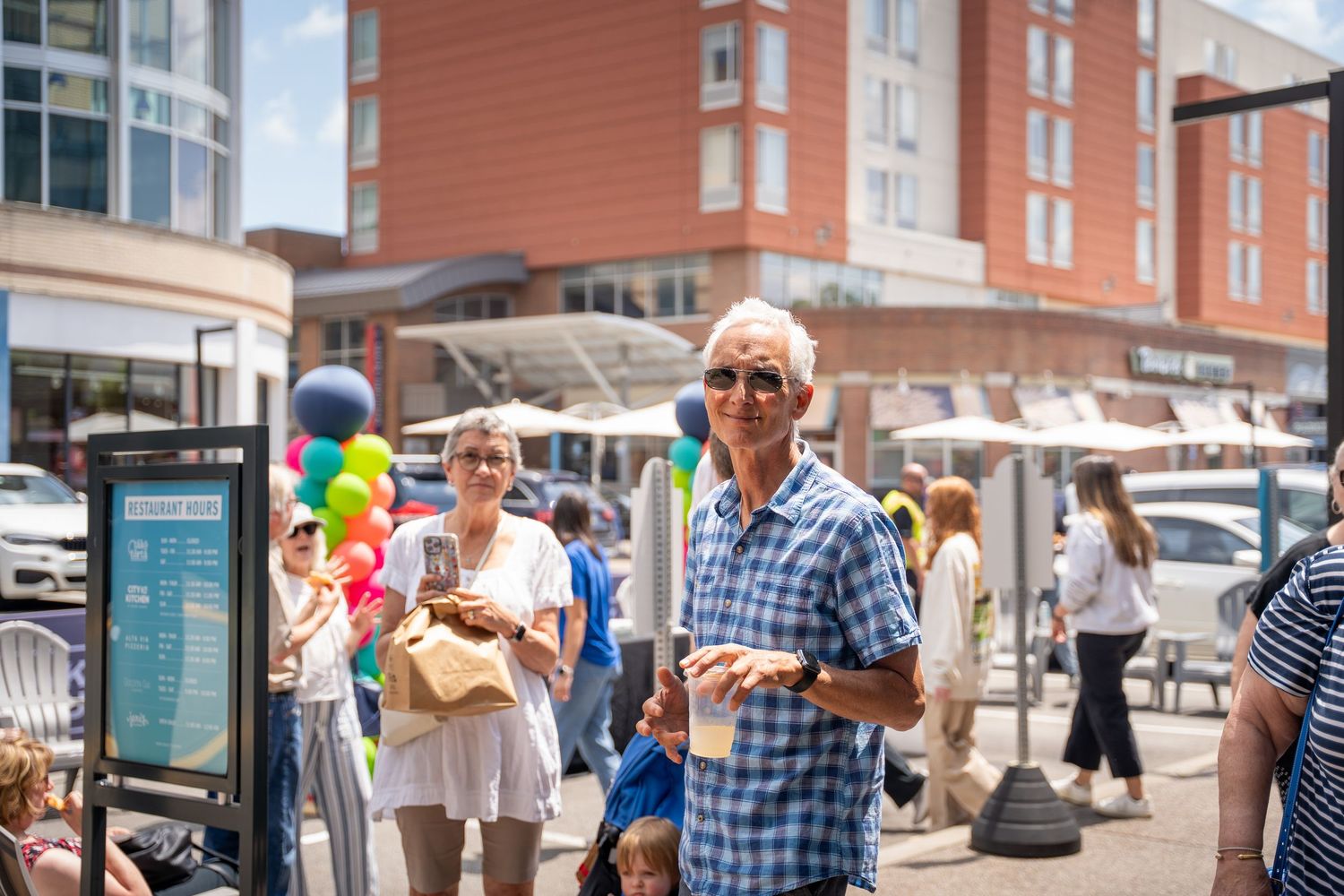People walk along a sunny sidewalk with colorful balloons and buildings in an urban setting.