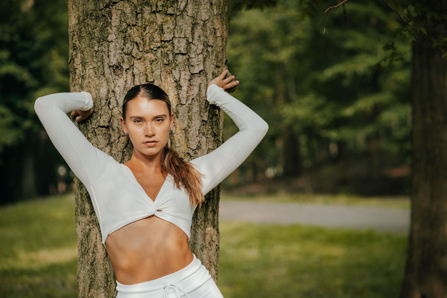 A person in a white long sleeve top poses against a tree trunk in an outdoor nature setting during golden hour.