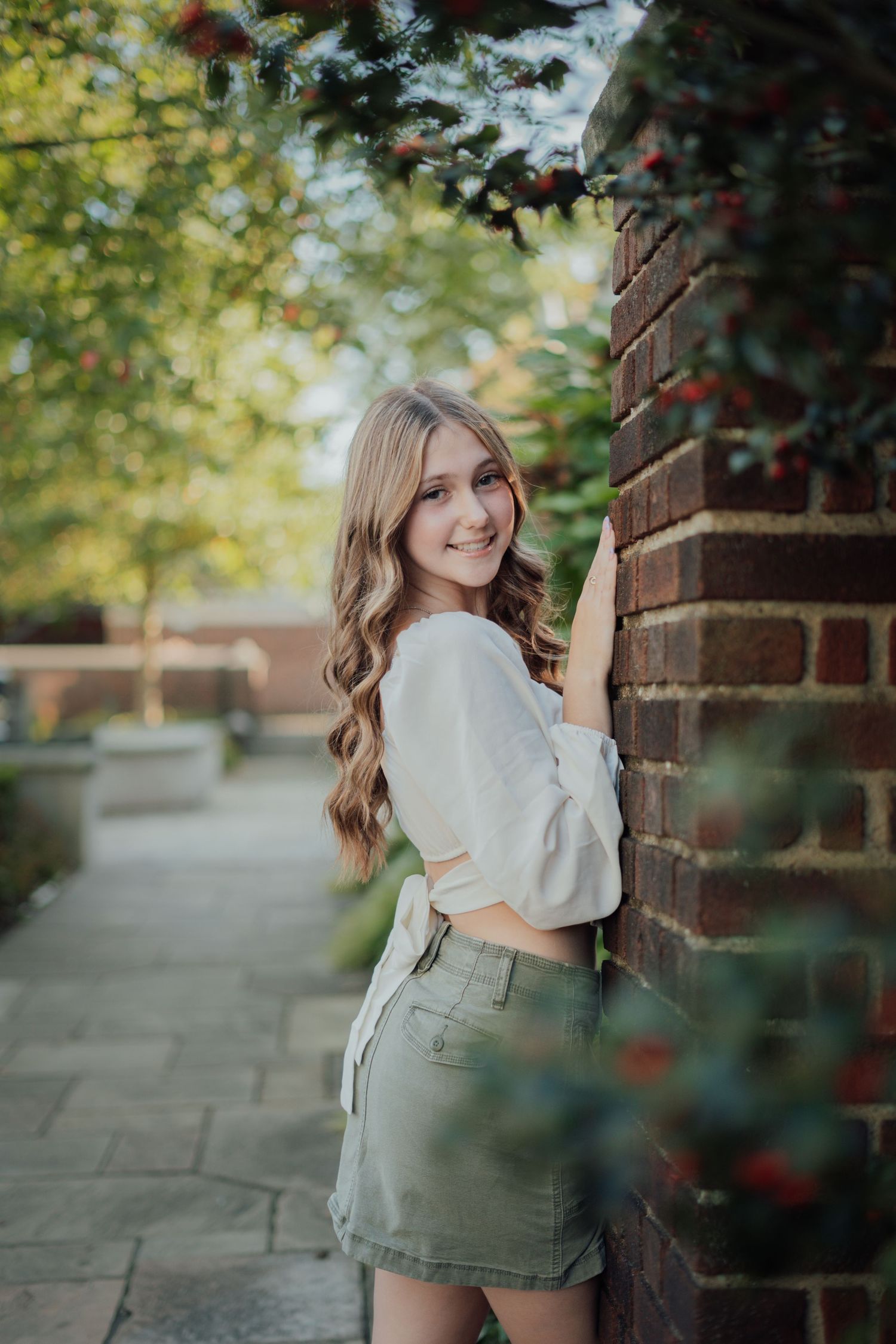 Portrait photo taken outdoors showing a casual pose against a brick wall with soft bokeh and greenery in background.