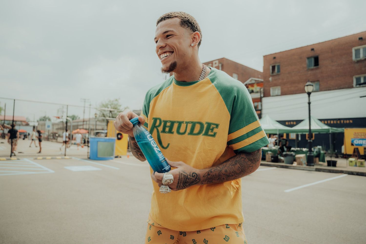 Athletic person wearing yellow and green RHODE sweatshirt smiles while holding water bottle in outdoor urban setting.