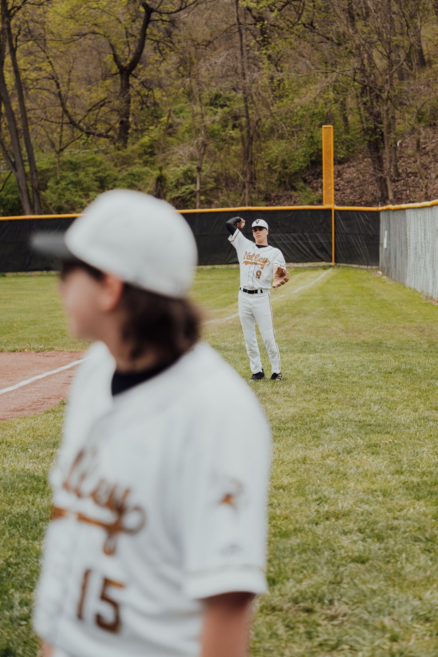 Baseball players in white uniforms practicing on a grass field with outfield fencing visible.