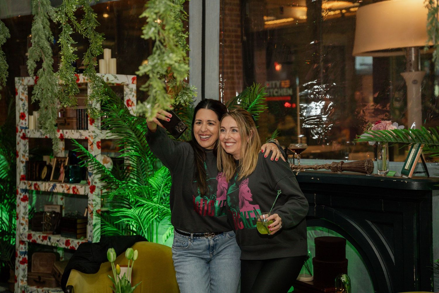 People mingle at indoor gathering space with exposed brick walls and hanging tropical plants illuminated in green light.