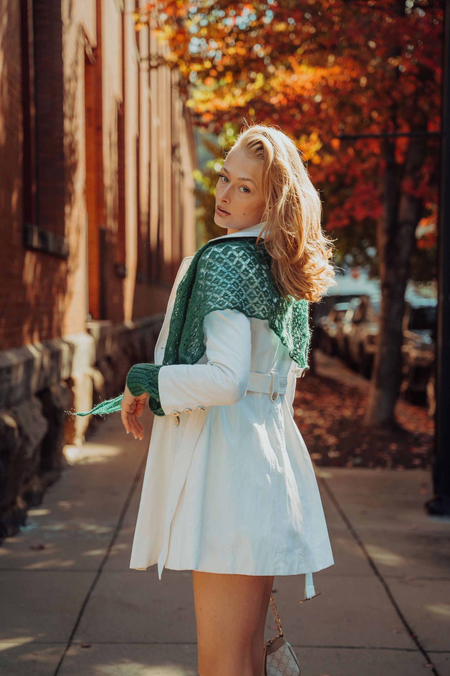 A woman in a white dress and green cardigan poses on an autumn sidewalk with colorful fall foliage in the background.