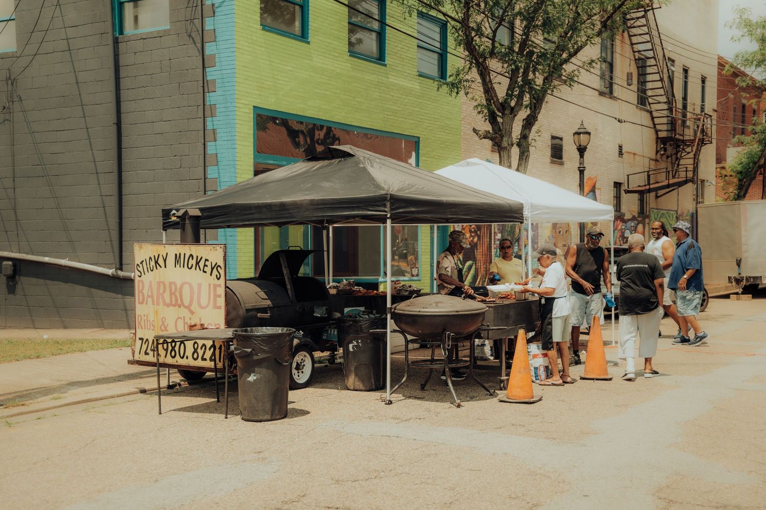 People wait in line at an outdoor food vendor with a white umbrella tent on a sunny city street corner.