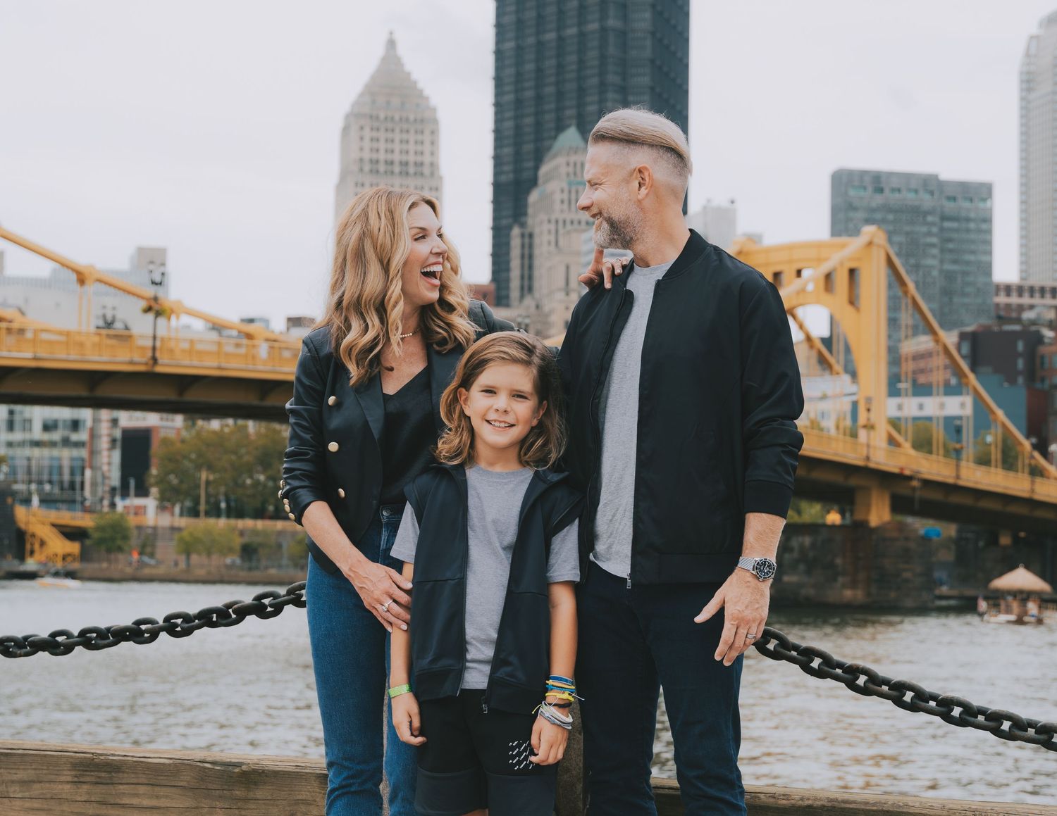 Couple laughs together during romantic urban engagement photo session along Pittsburgh riverfront near iconic bridges.