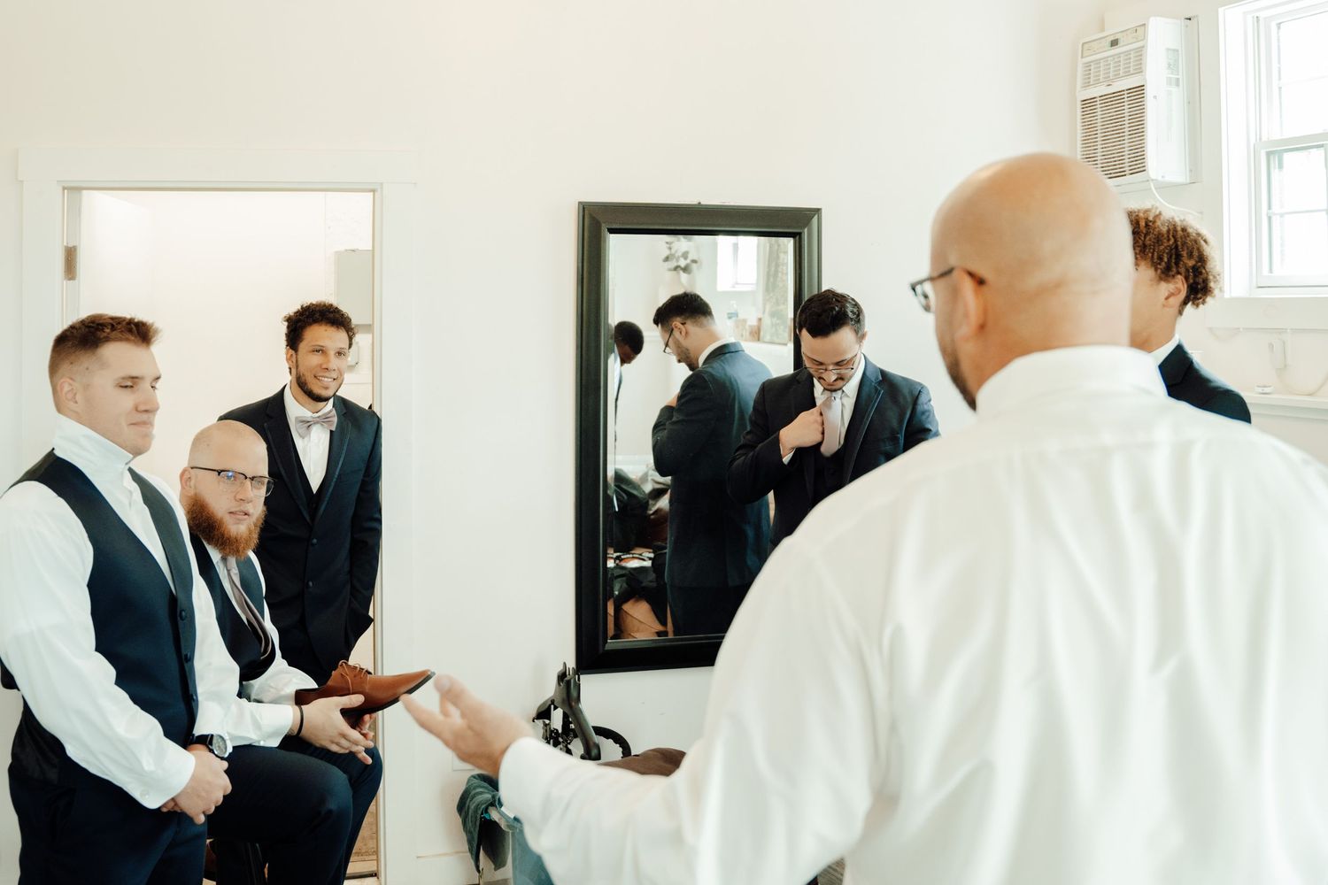 Groomsmen getting ready in bright room with window light while others reflect in mirror.