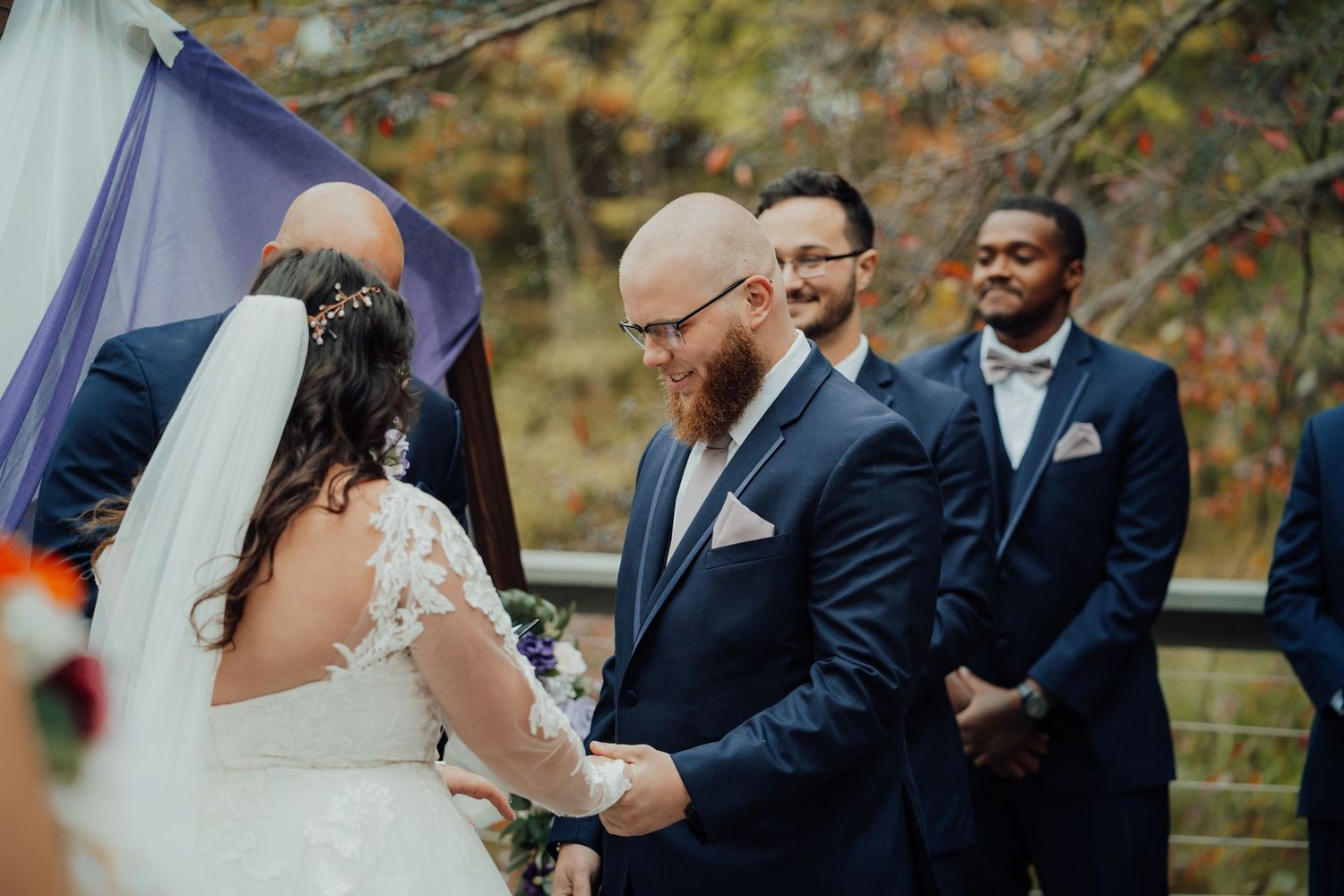 Couple holding hands during outdoor wedding ceremony with navy backdrop and fall foliage.