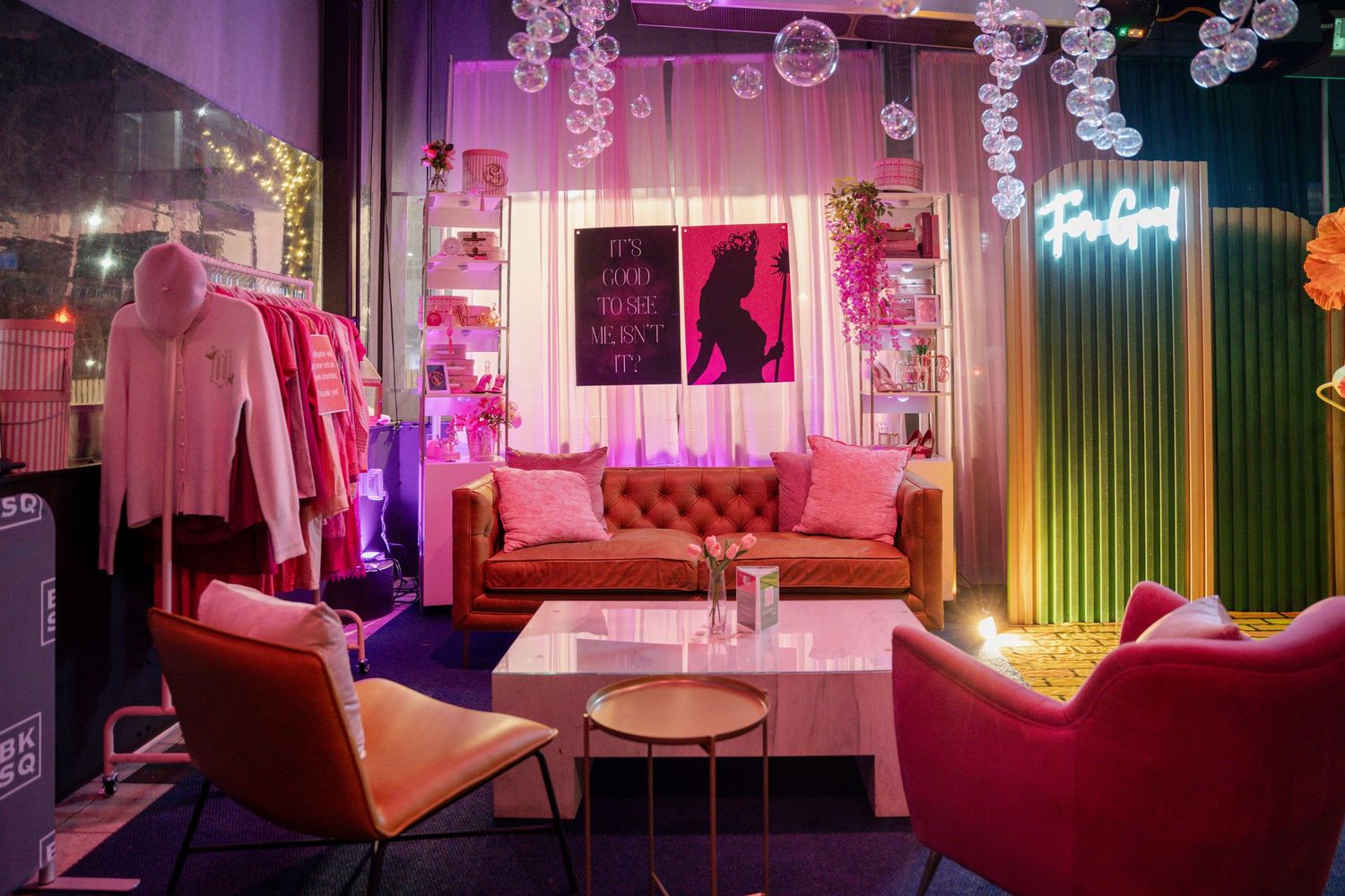 Stylish event space featuring pink velvet sofa, bubble decorations, and neon-lit ambiance with clothing displays.