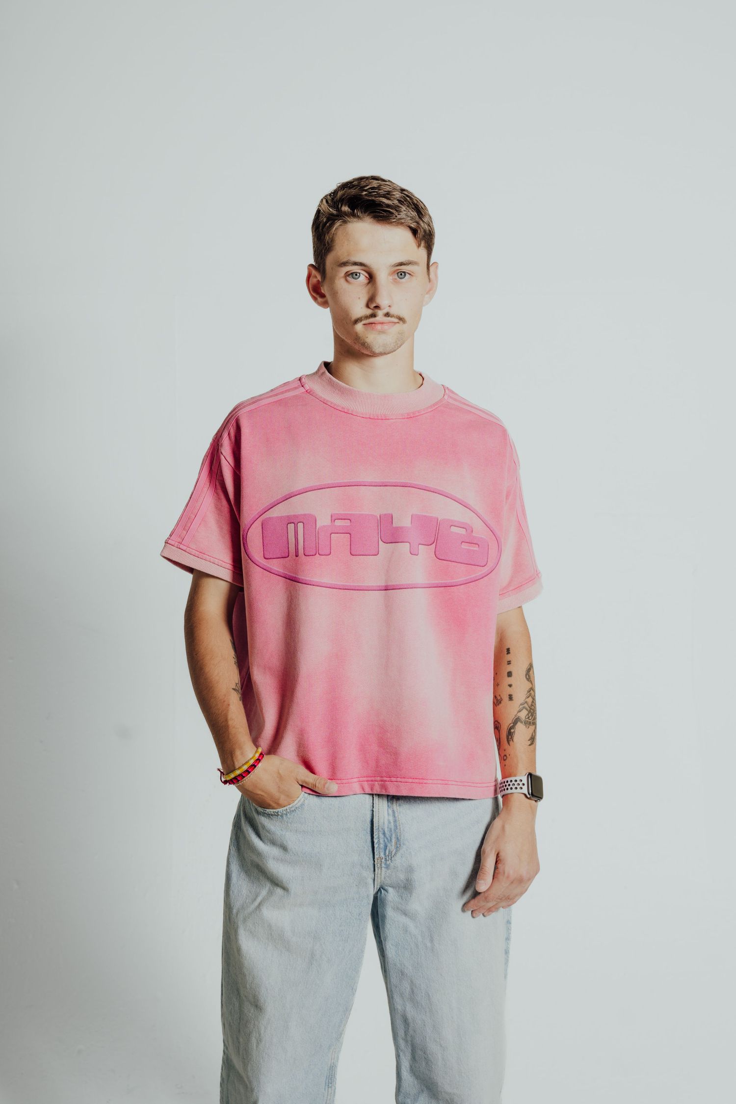 Pink retro-style t-shirt with oval logo worn with light wash jeans against white background.